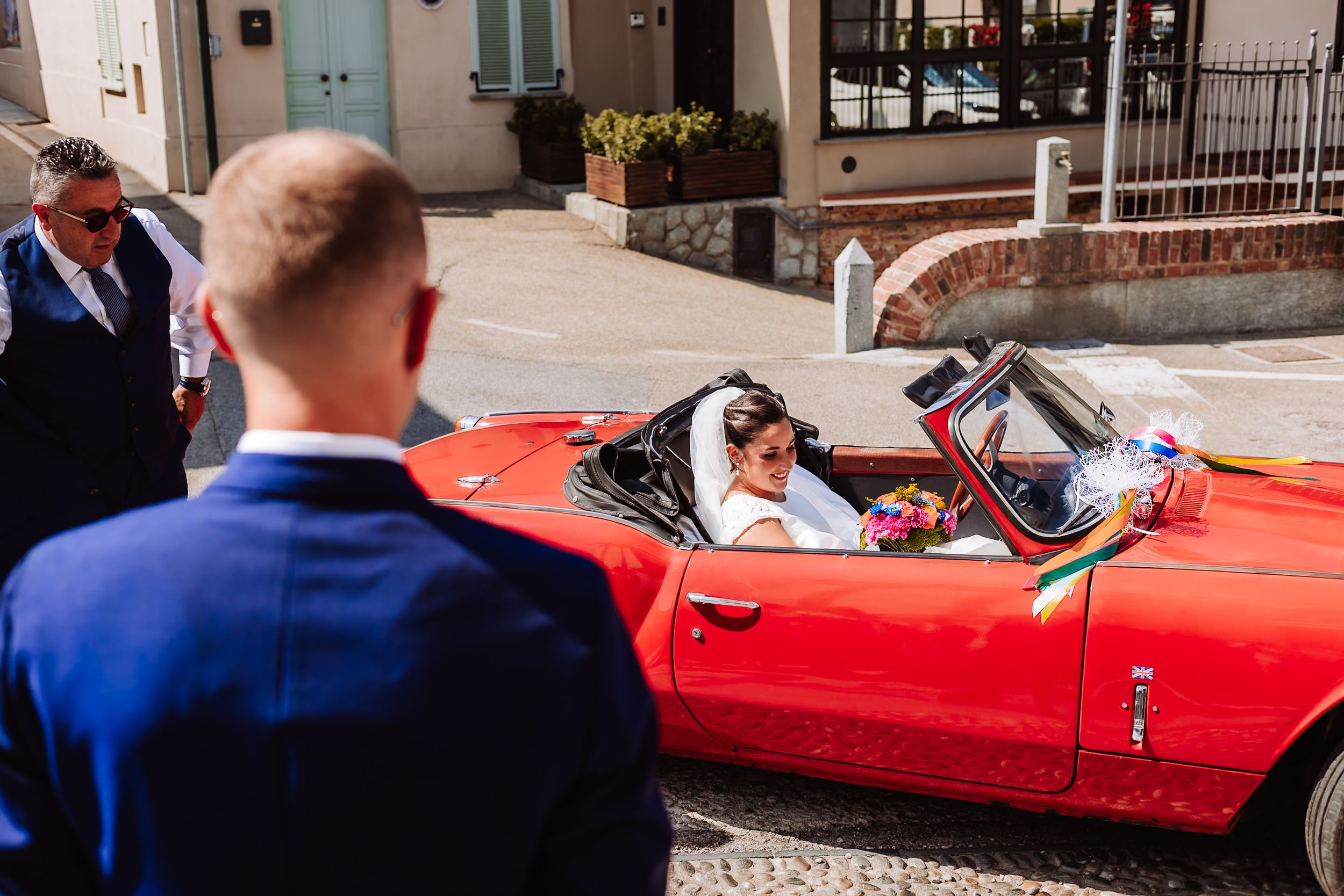 Bride in white dress seated in a red convertible classic car, with groom in blue suit standing nearby on a sunny day.