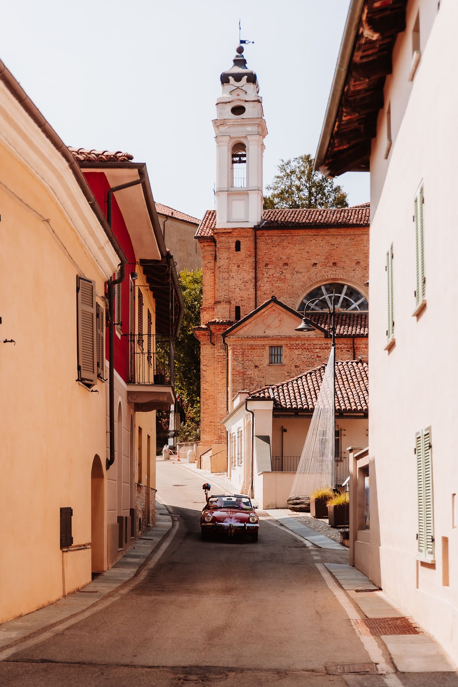 A vintage red convertible car drives through a narrow Italian village street with a white church bell tower in the background.