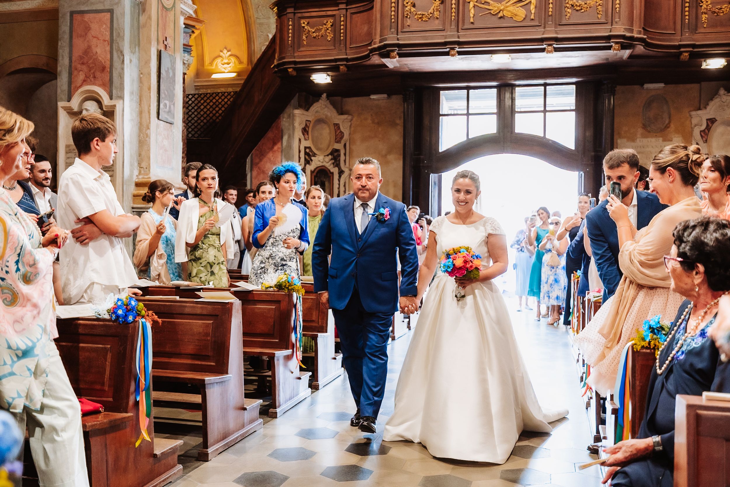 Bride and groom walking down church aisle during wedding ceremony, surrounded by guests, colorful bouquet.