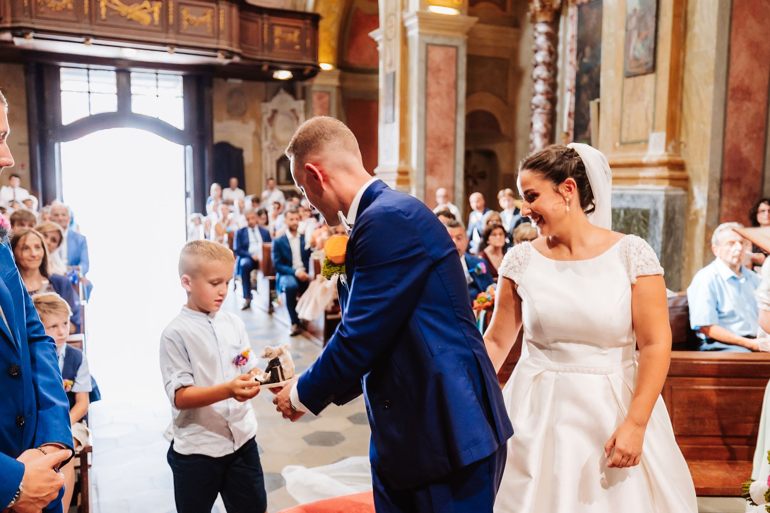 Groom in blue suit exchanges rings with bride in white dress as young boy assists during church wedding ceremony.