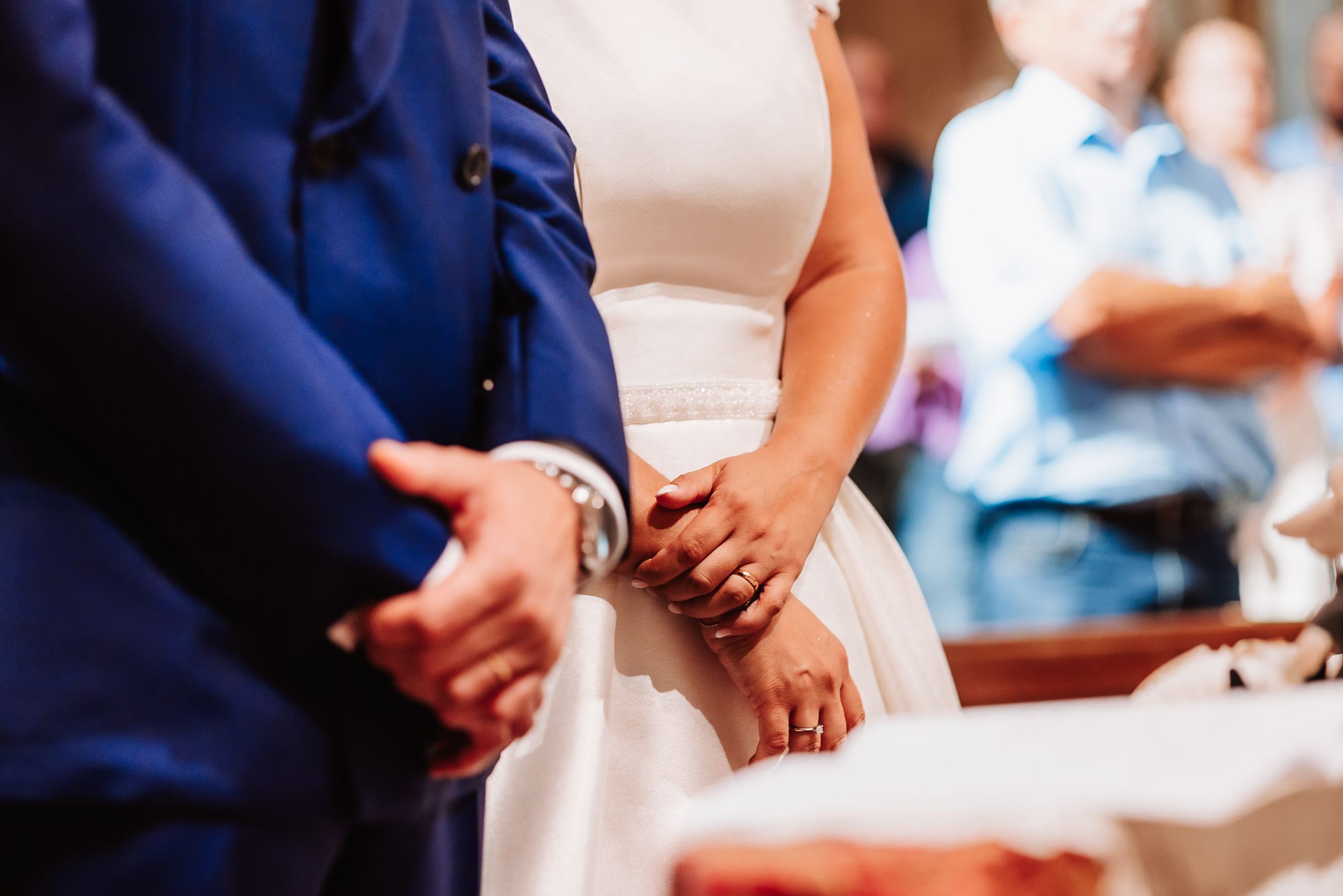 Close-up of bride and groom holding hands during wedding ceremony, wearing rings, bride in white dress.