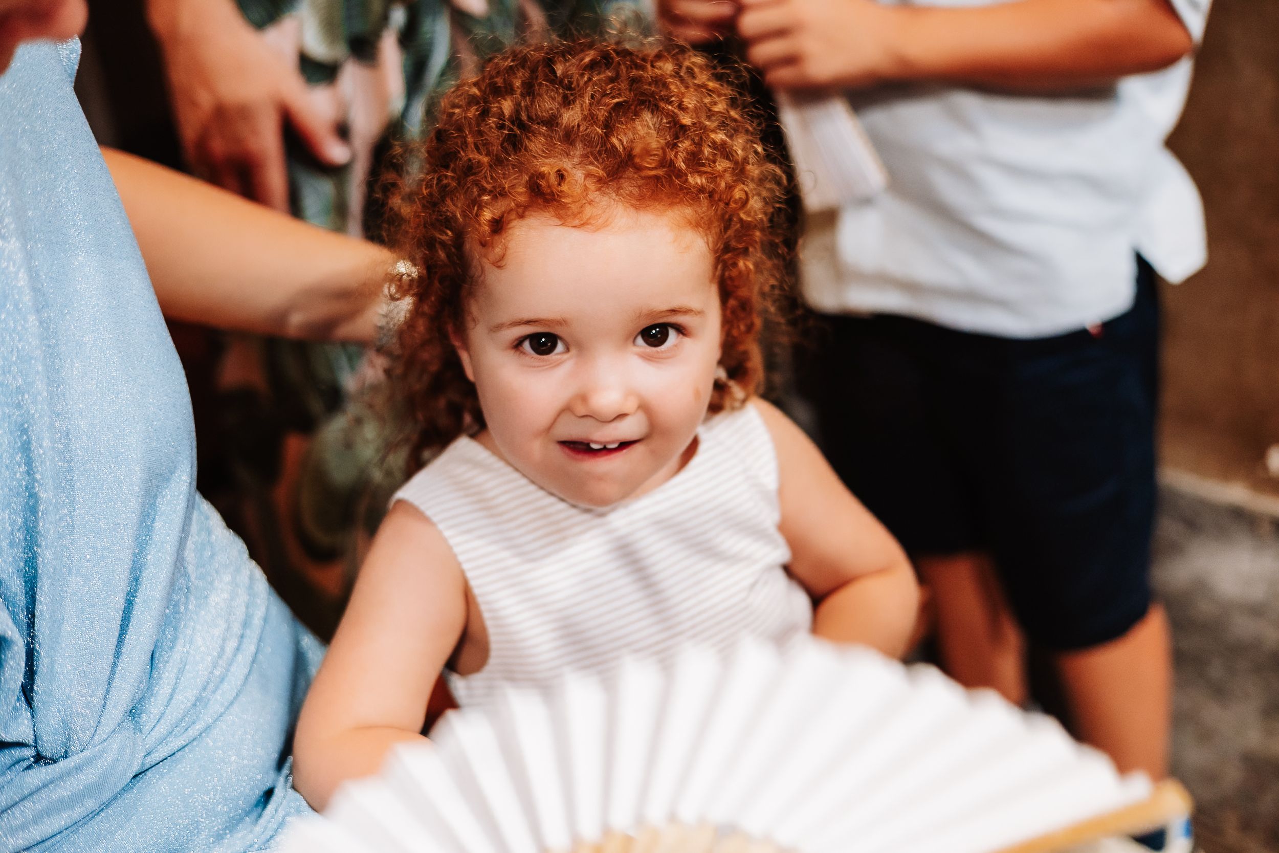 Adorable curly red-haired toddler in white dress holding a hand fan, smiling at camera outdoors.