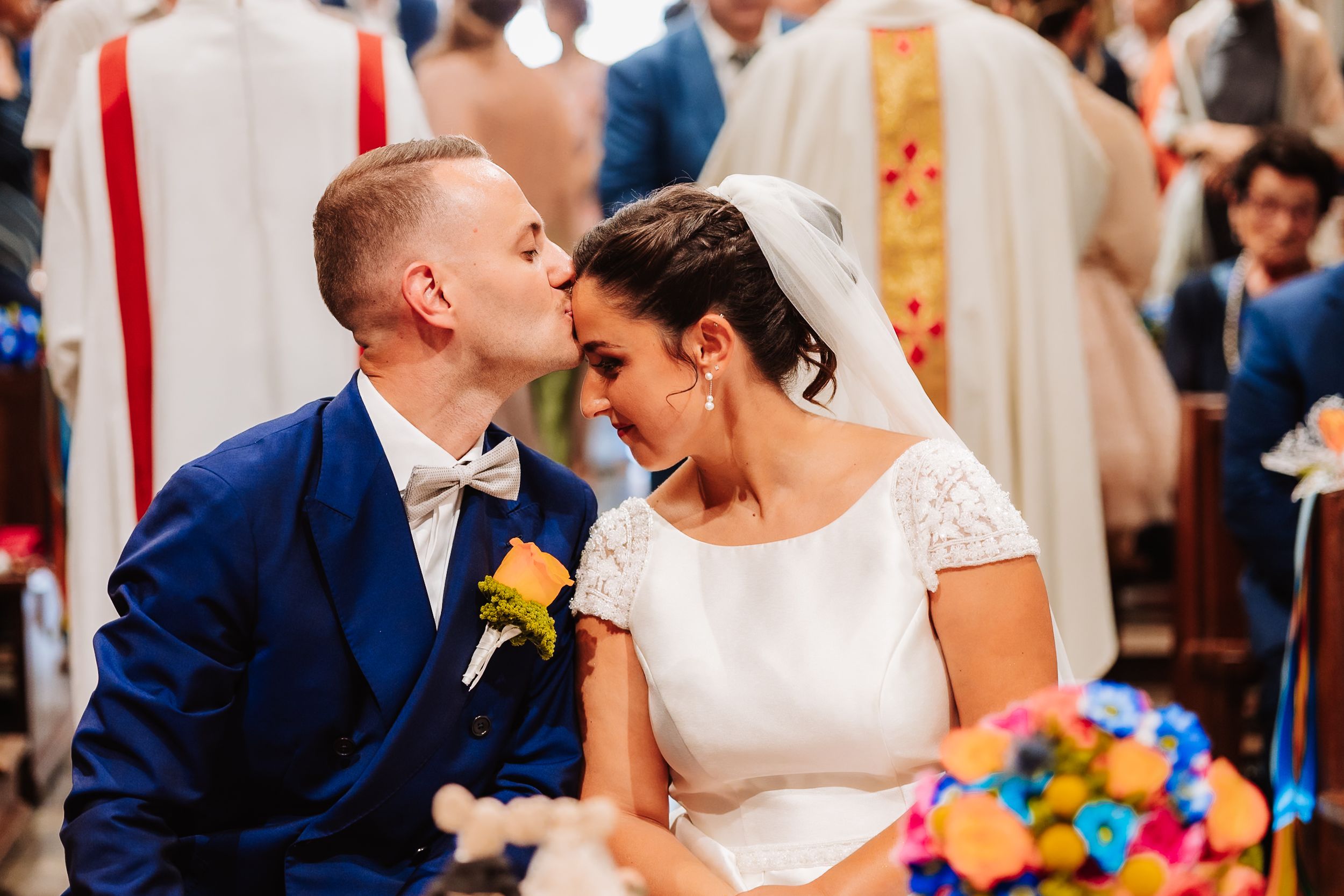 Bride and groom sharing a kiss at church altar, groom in blue suit with yellow boutonniere, bride in white dress with veil.
