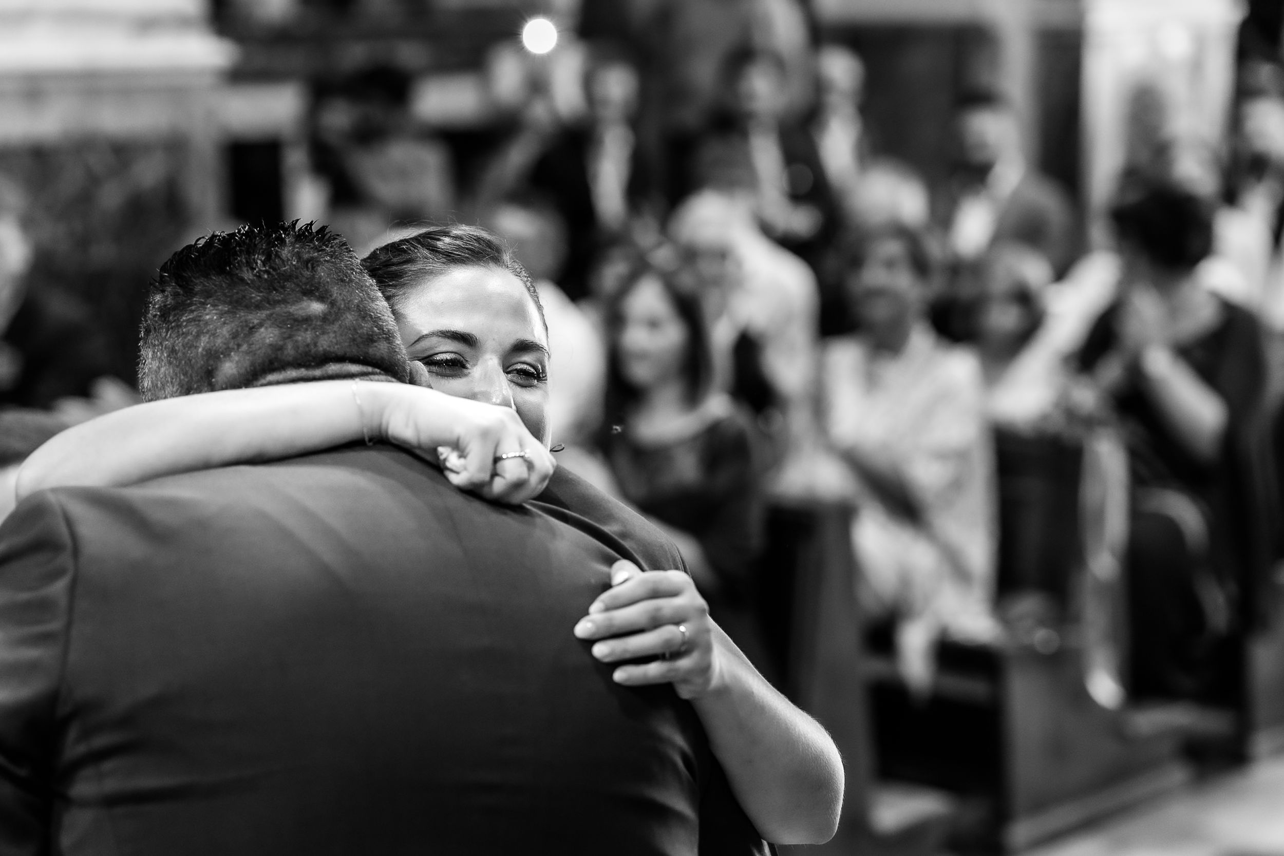 Black and white photo of a couple sharing an emotional hug at a wedding ceremony with guests seated in background.
