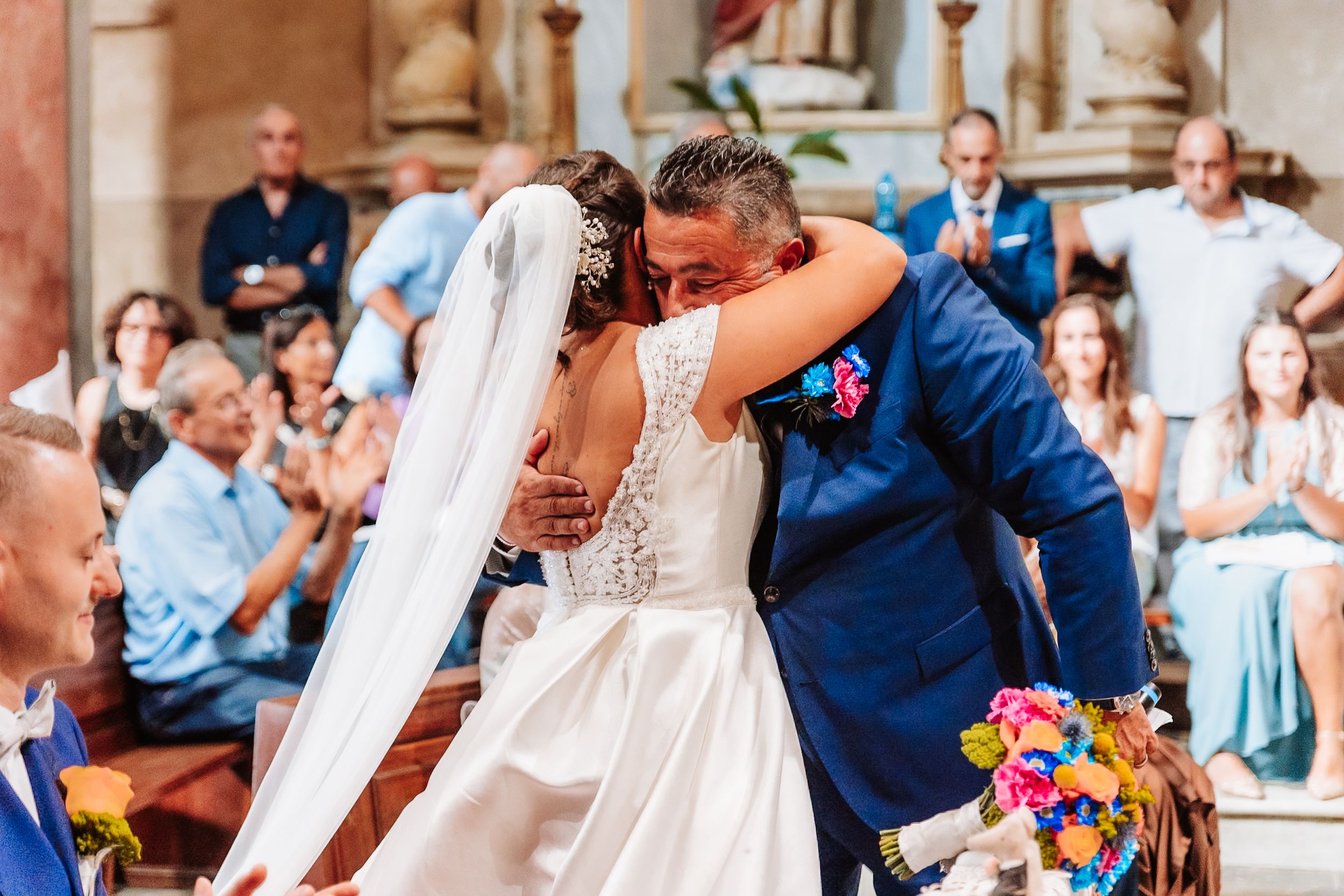 Newlyweds sharing a heartfelt embrace at the altar surrounded by applauding wedding guests in a church.