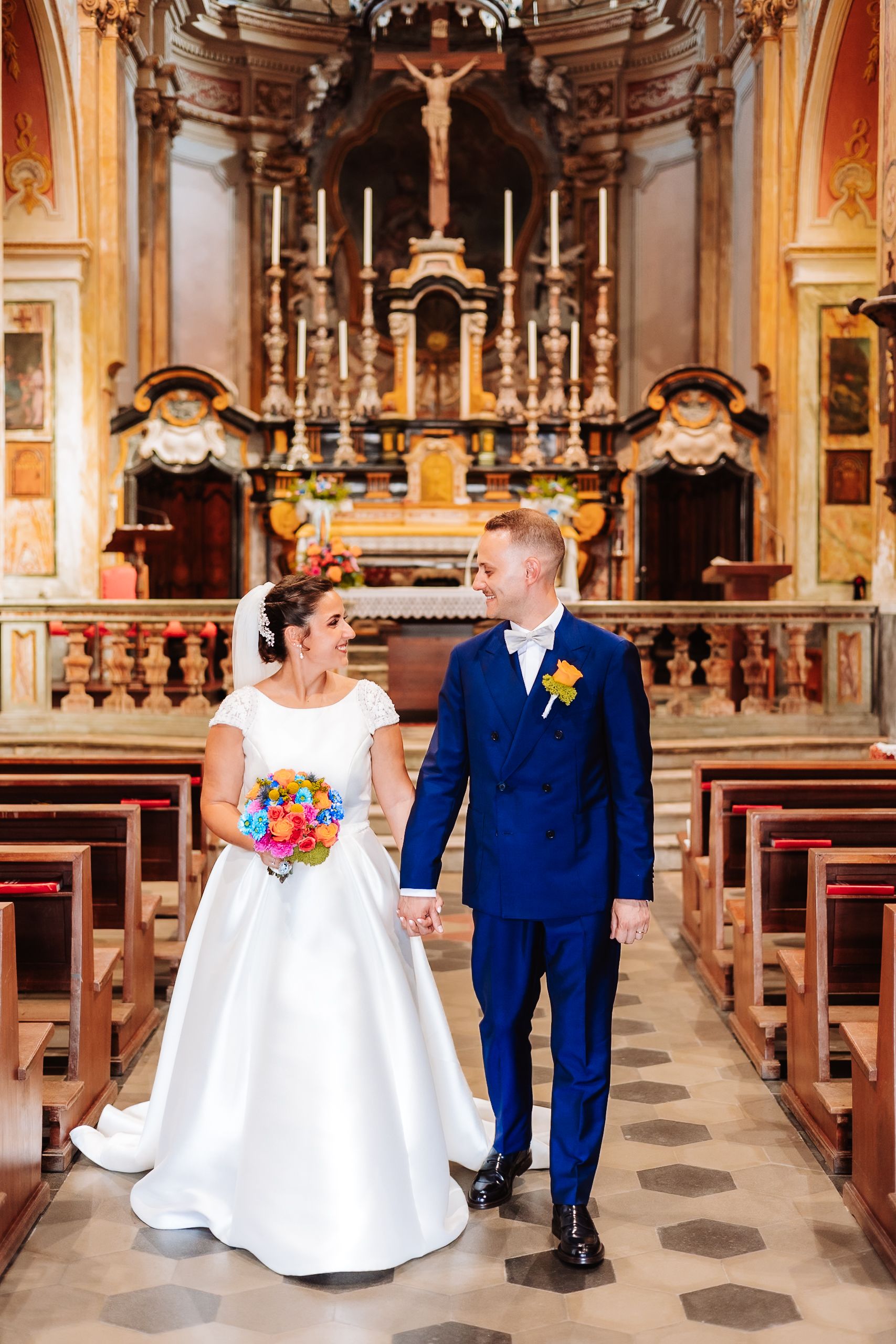 Bride and groom walking hand in hand down the aisle of an ornate church, bride holding colorful bouquet.