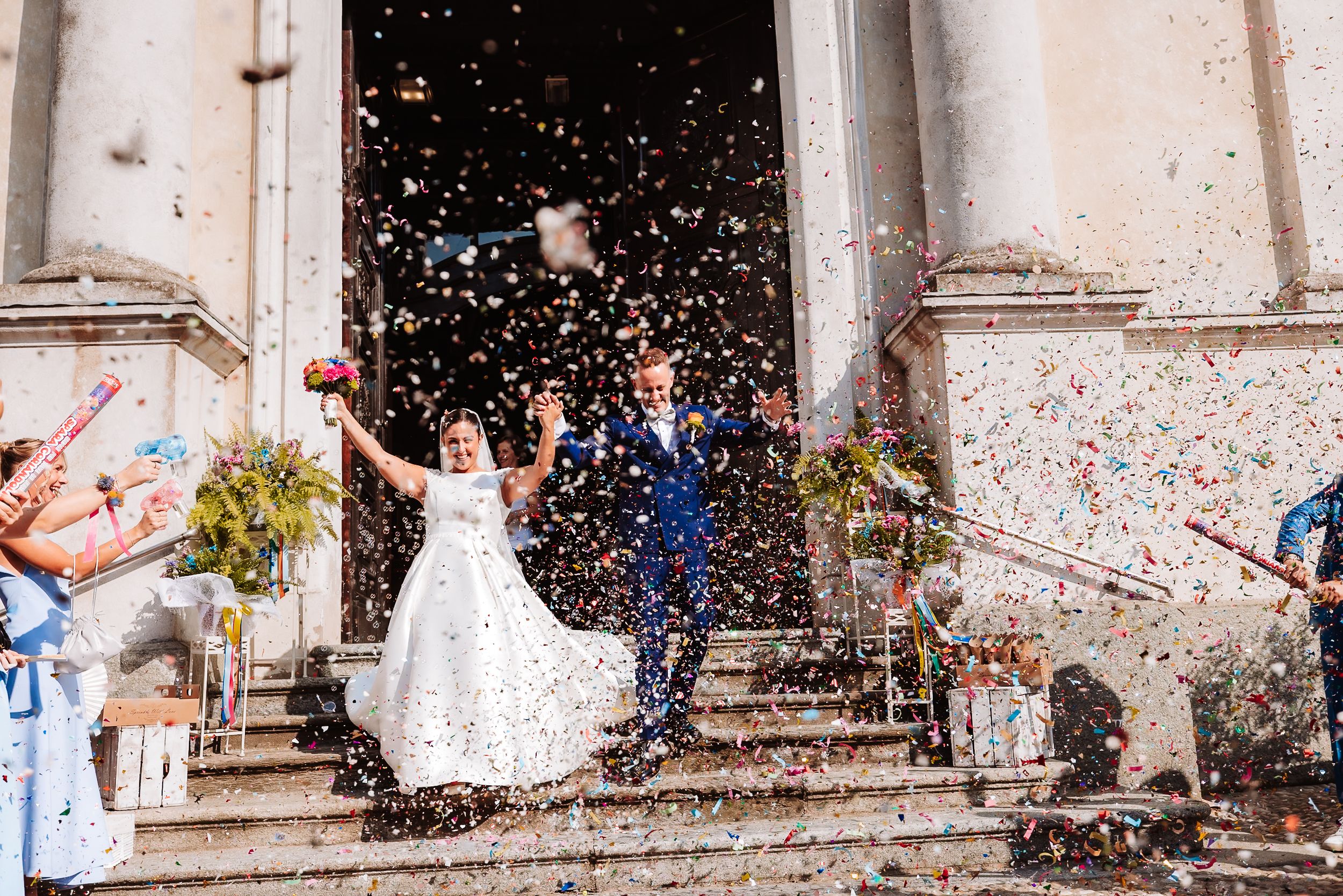 Joyful newlyweds exit church amid colorful confetti shower, celebrating their wedding day.