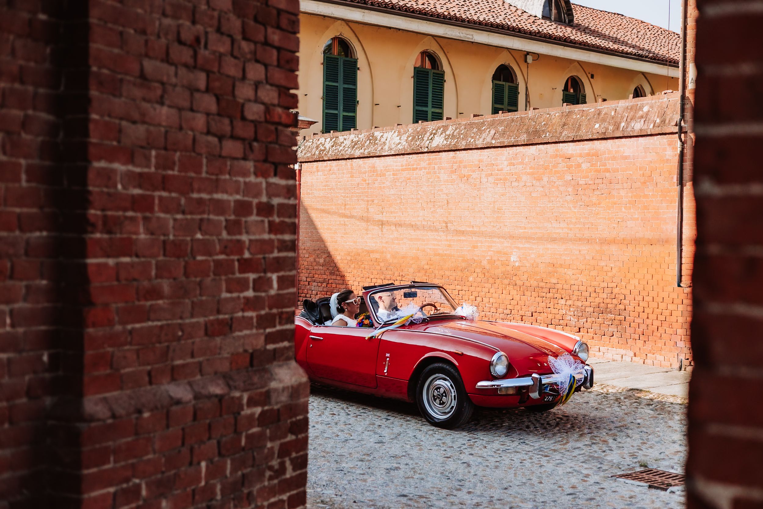 A classic red vintage convertible sports car parked on cobblestones between rustic brick walls in an Italian town.