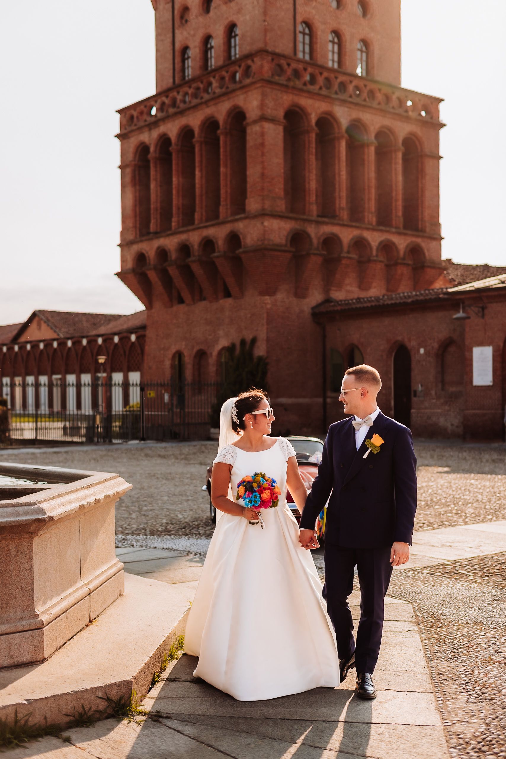 Bride and groom walk hand-in-hand near a historic red brick tower on their wedding day.