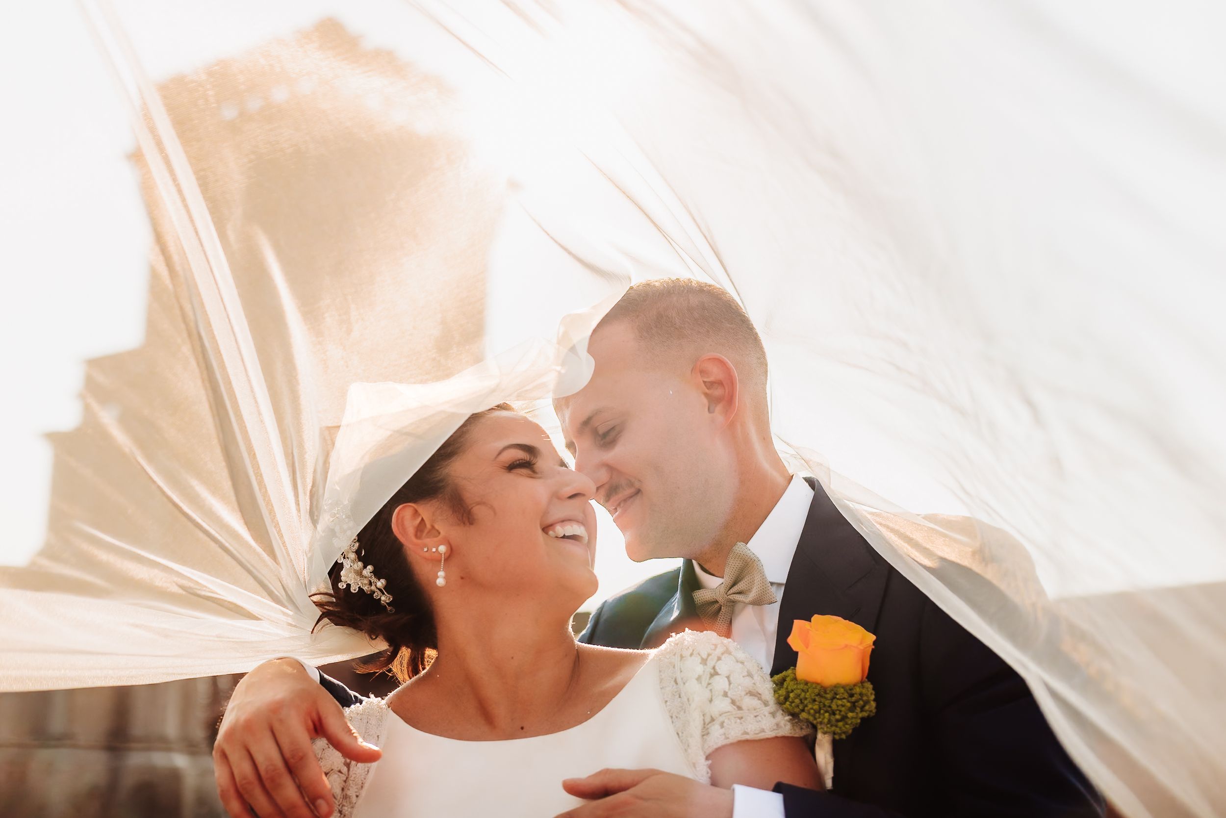 Bride and groom share romantic moment under flowing veil with golden sunlight backdrop on wedding day.