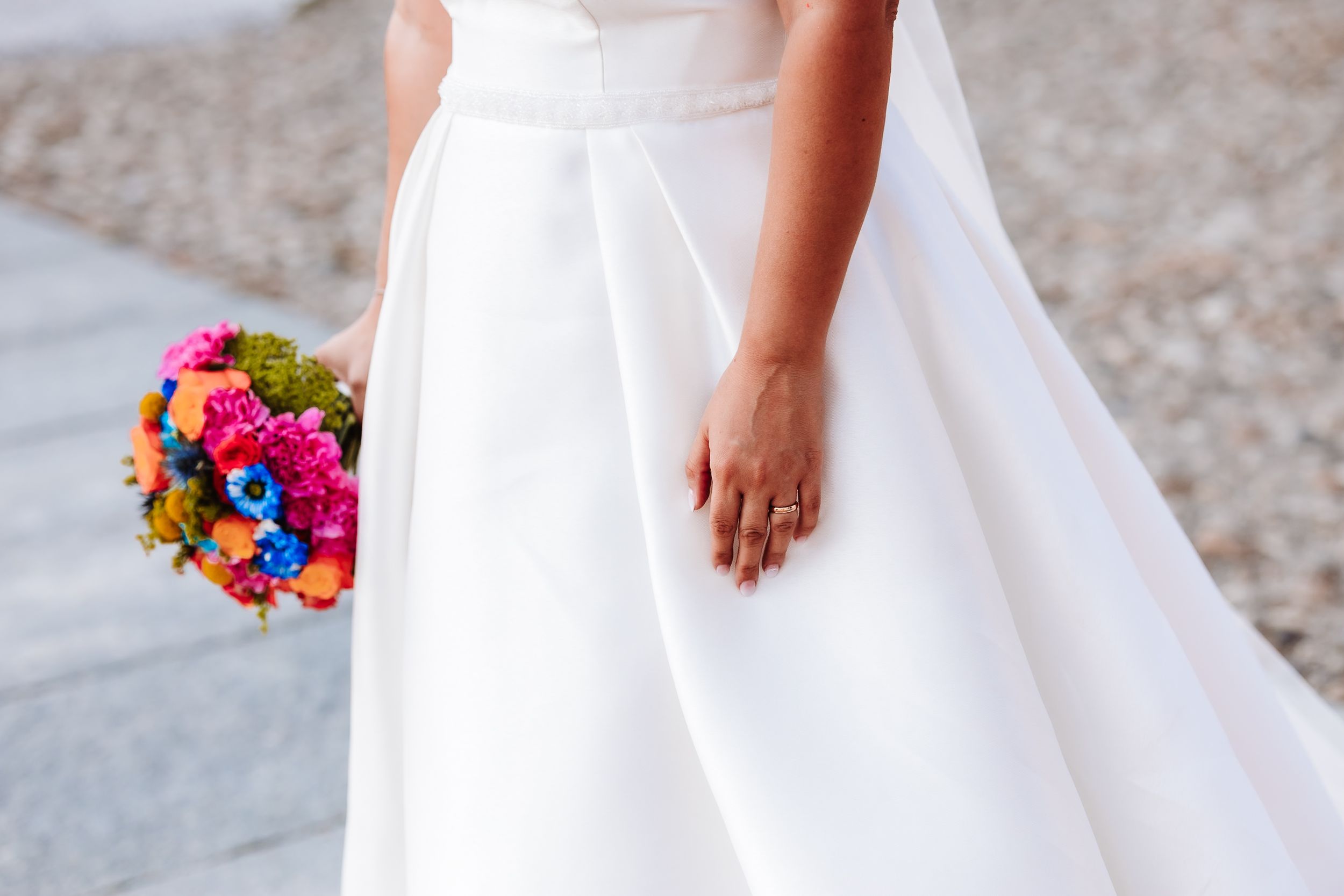 Bride in white wedding dress holding colorful bouquet of pink, blue, and orange flowers at outdoor ceremony.