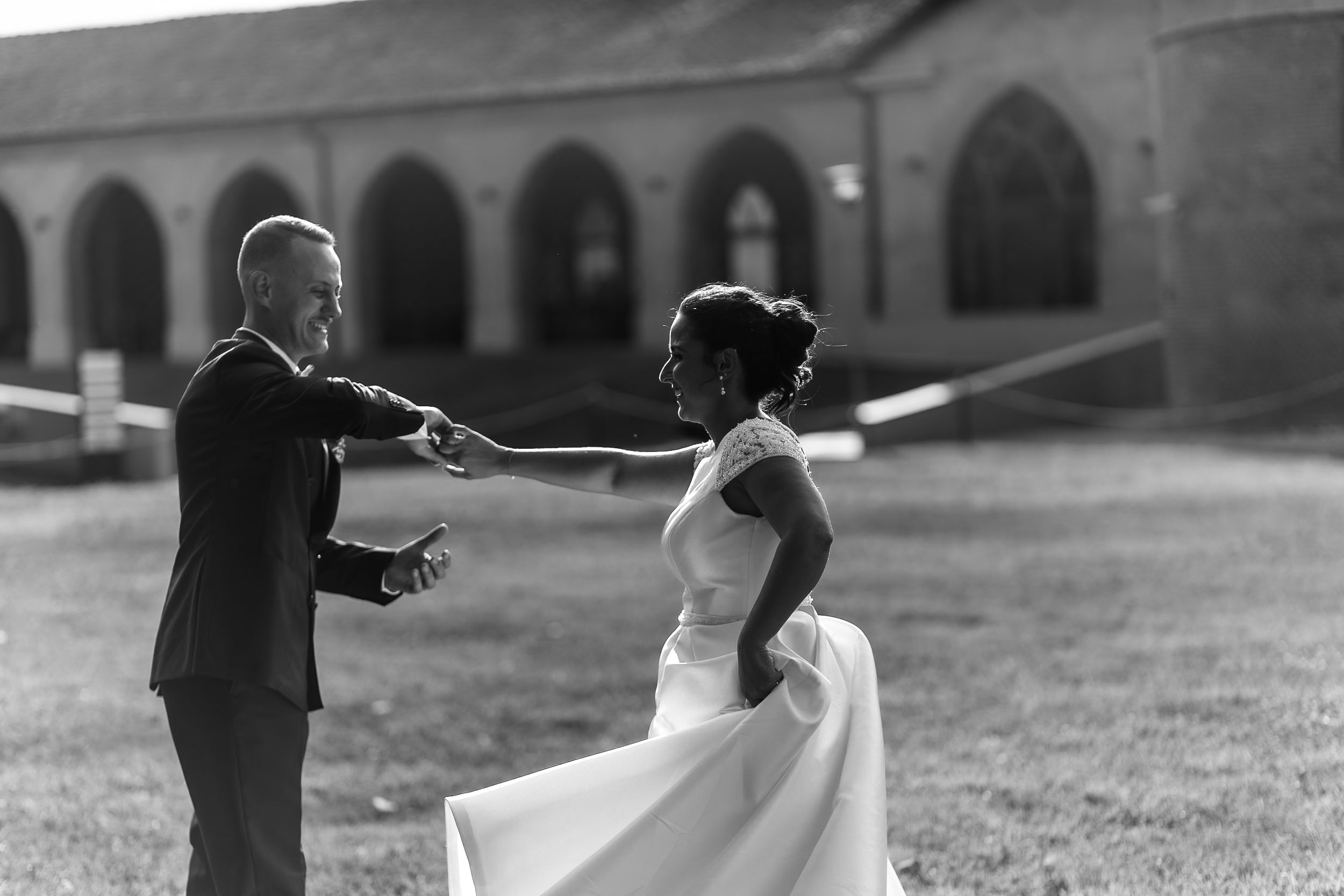 Black and white photo of a couple dancing outdoors at their wedding, with arched stone building in background.