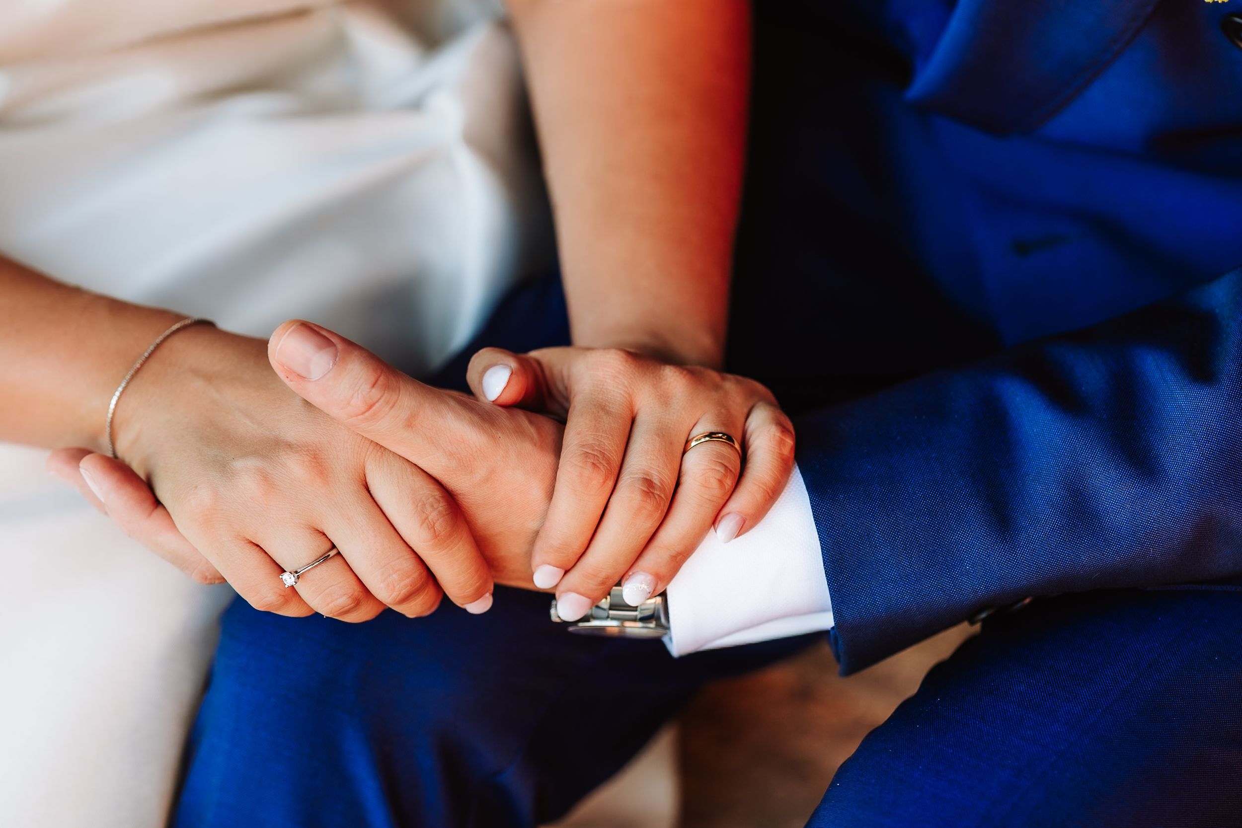 Close-up of a bride and groom holding hands, showing wedding rings, with the groom in a blue suit.