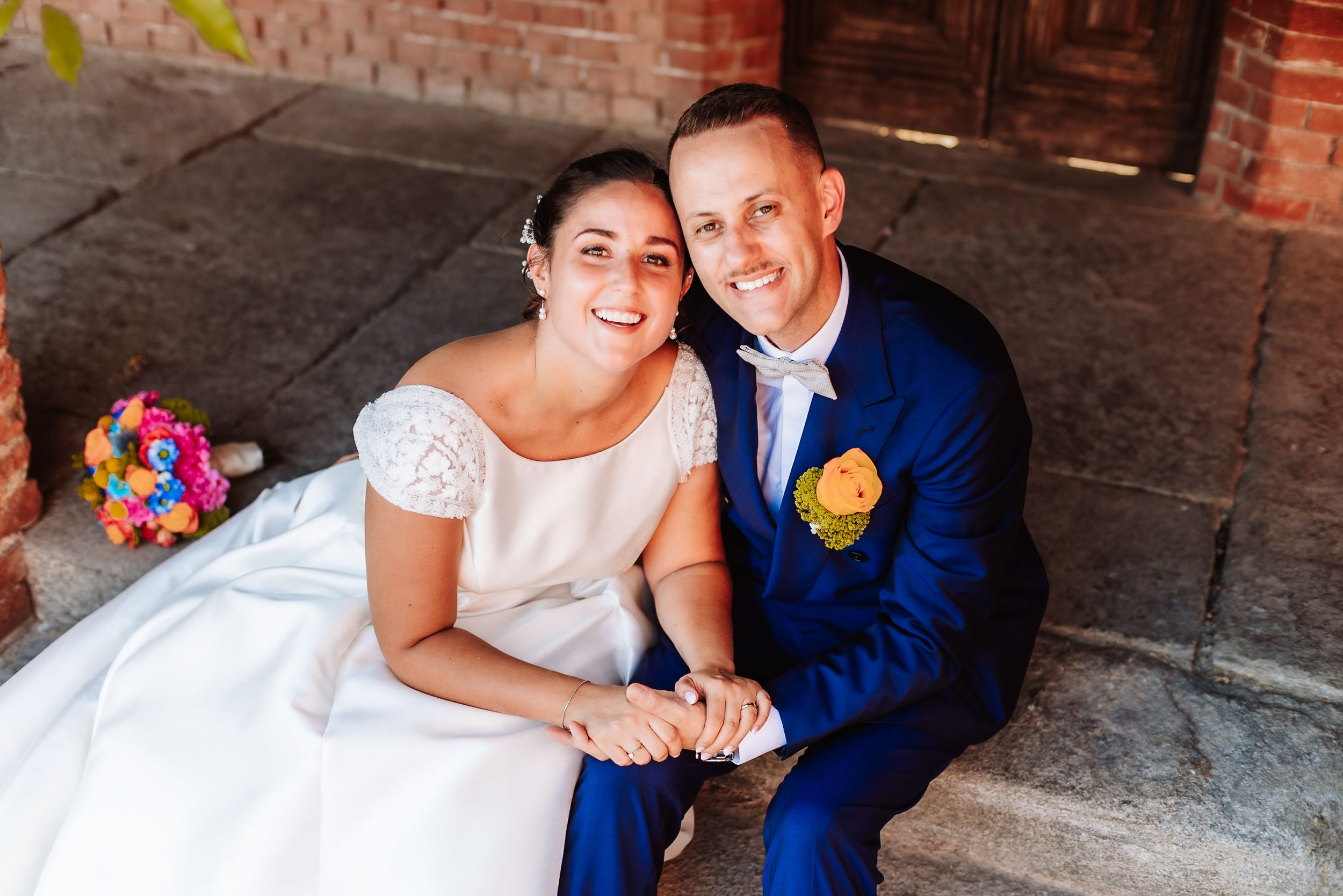 Happy newlywed couple sitting on steps, bride in white gown holding colorful bouquet, groom in blue suit with yellow boutonniere.