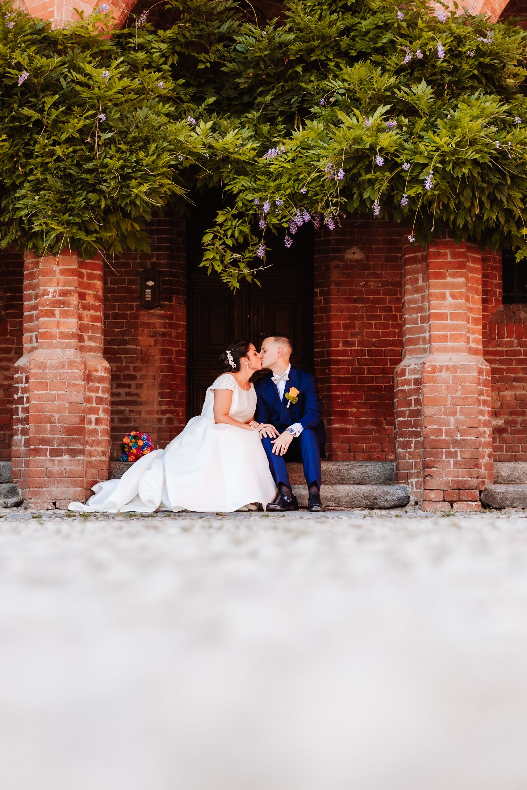 Bride and groom share a kiss seated before a red brick wall adorned with wisteria.