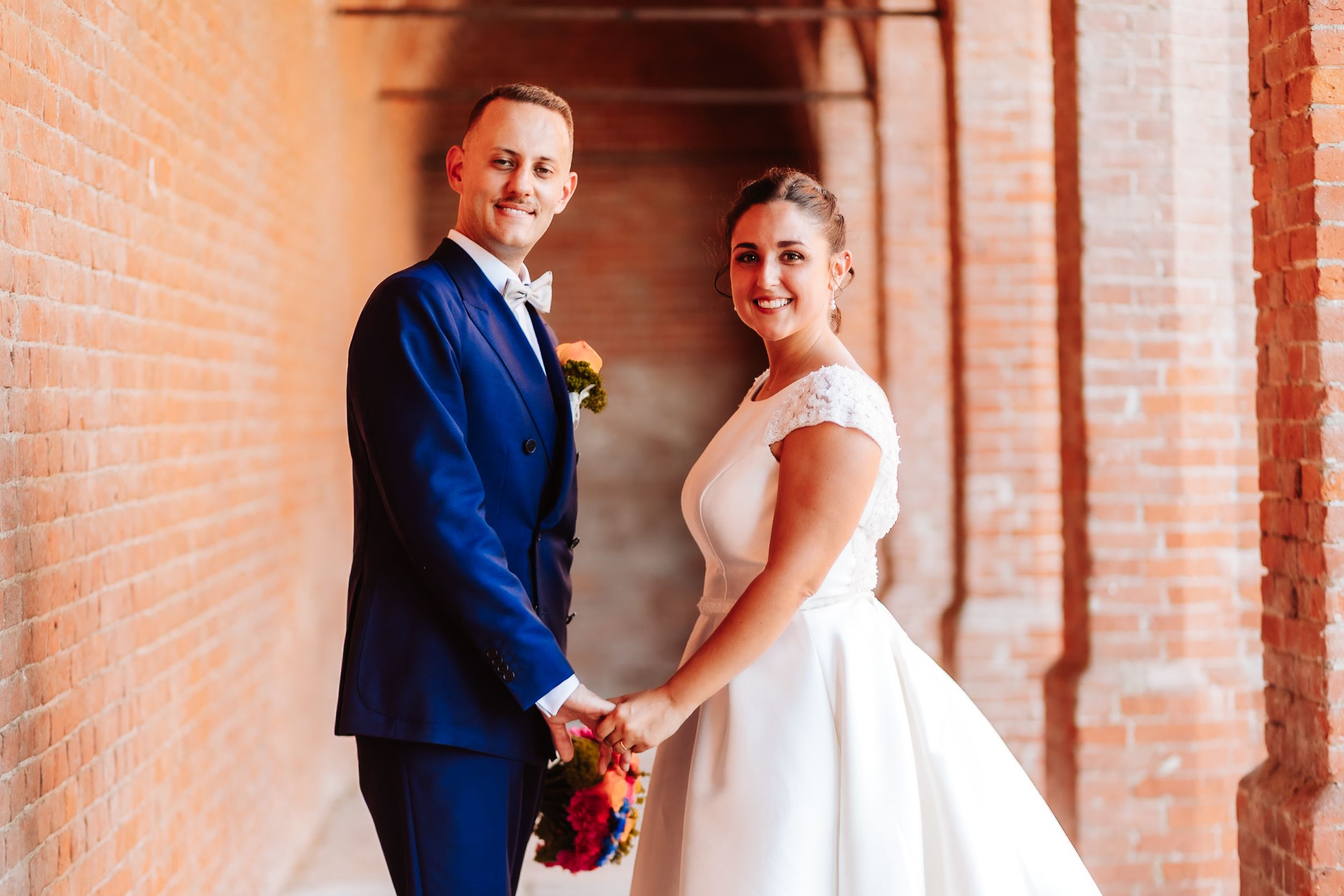 Newlyweds hold hands and smile in brick archway, groom in blue suit and bride in white ballgown.