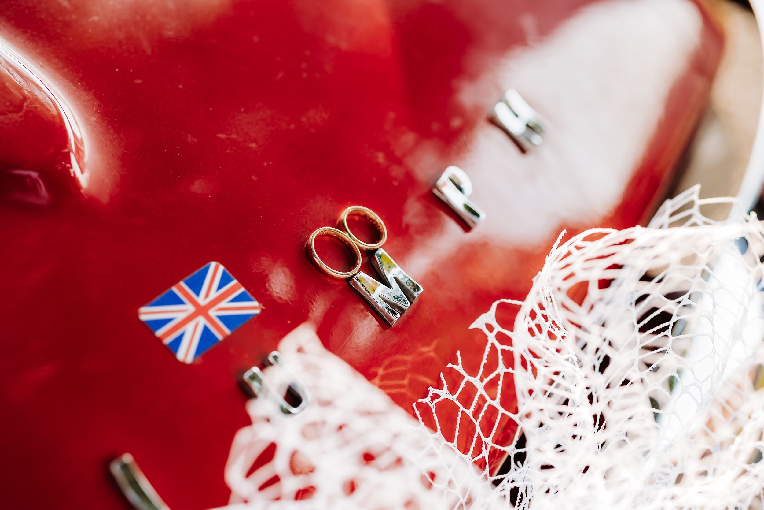 Close-up of wedding rings and small Union Jack flag on a shiny red surface with white lace detail.