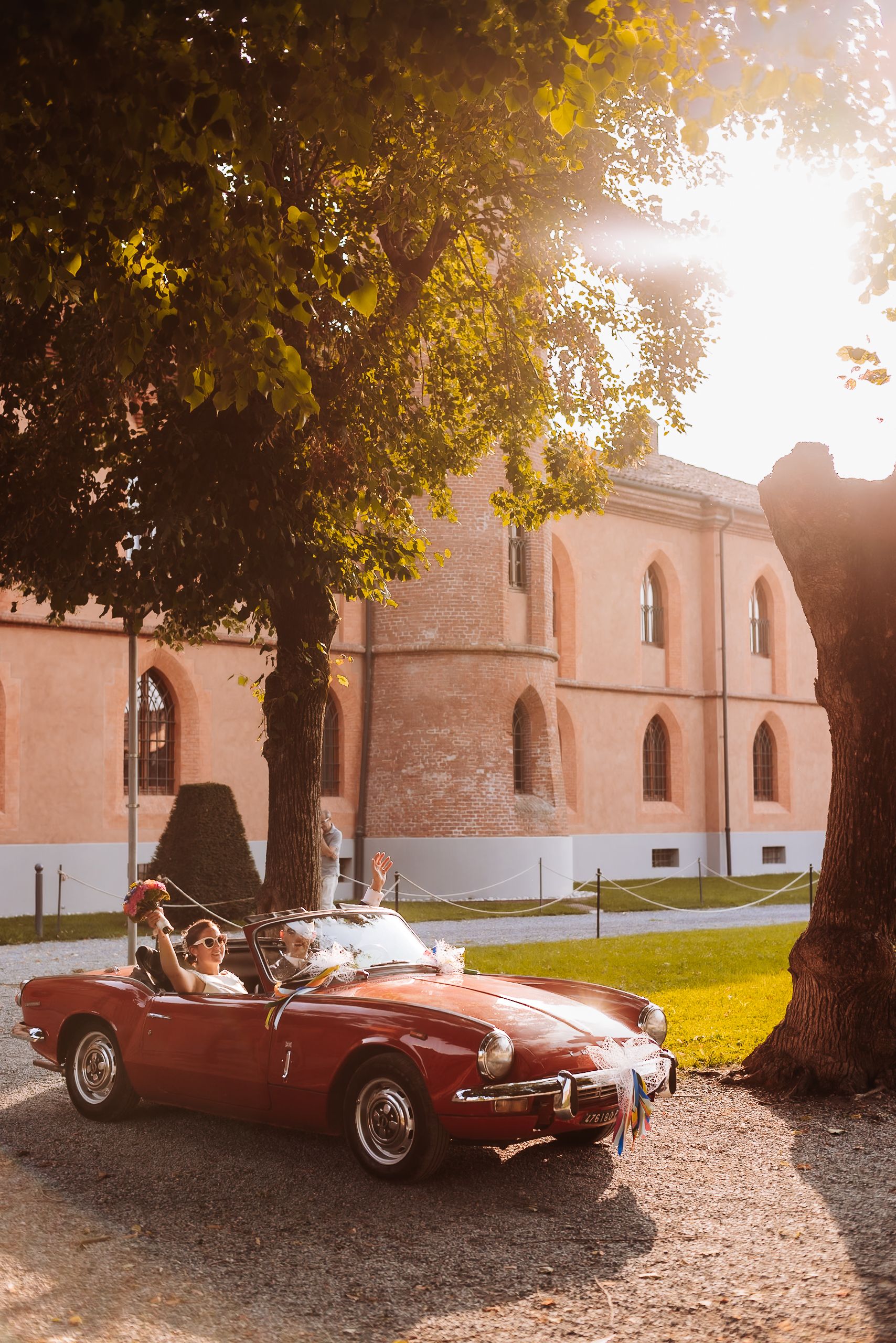 Couple riding in a vintage red convertible sports car near a historic brick building at golden hour.