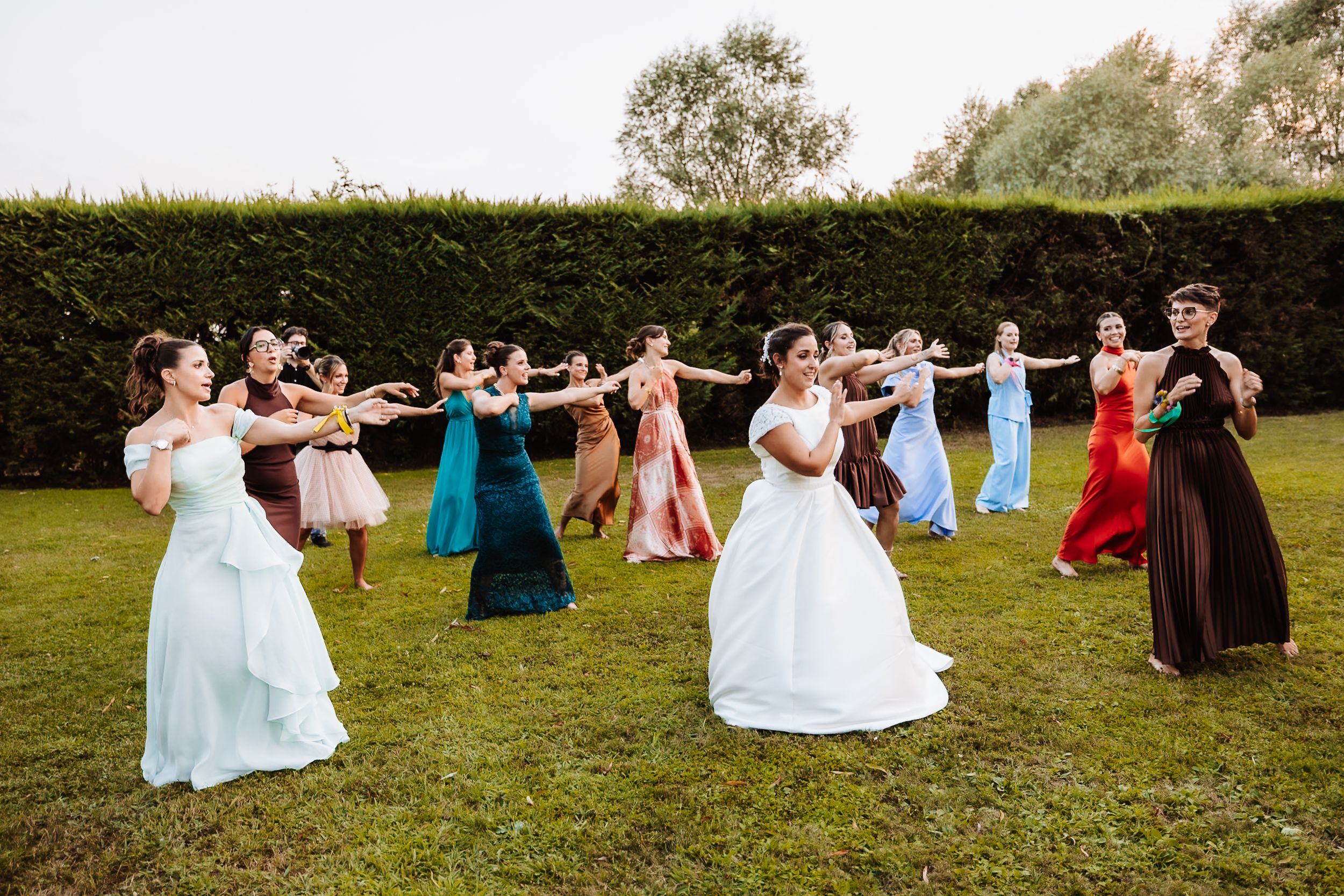 Bridesmaids in colorful gowns dance in a chain with the bride on a green lawn at an outdoor wedding.
