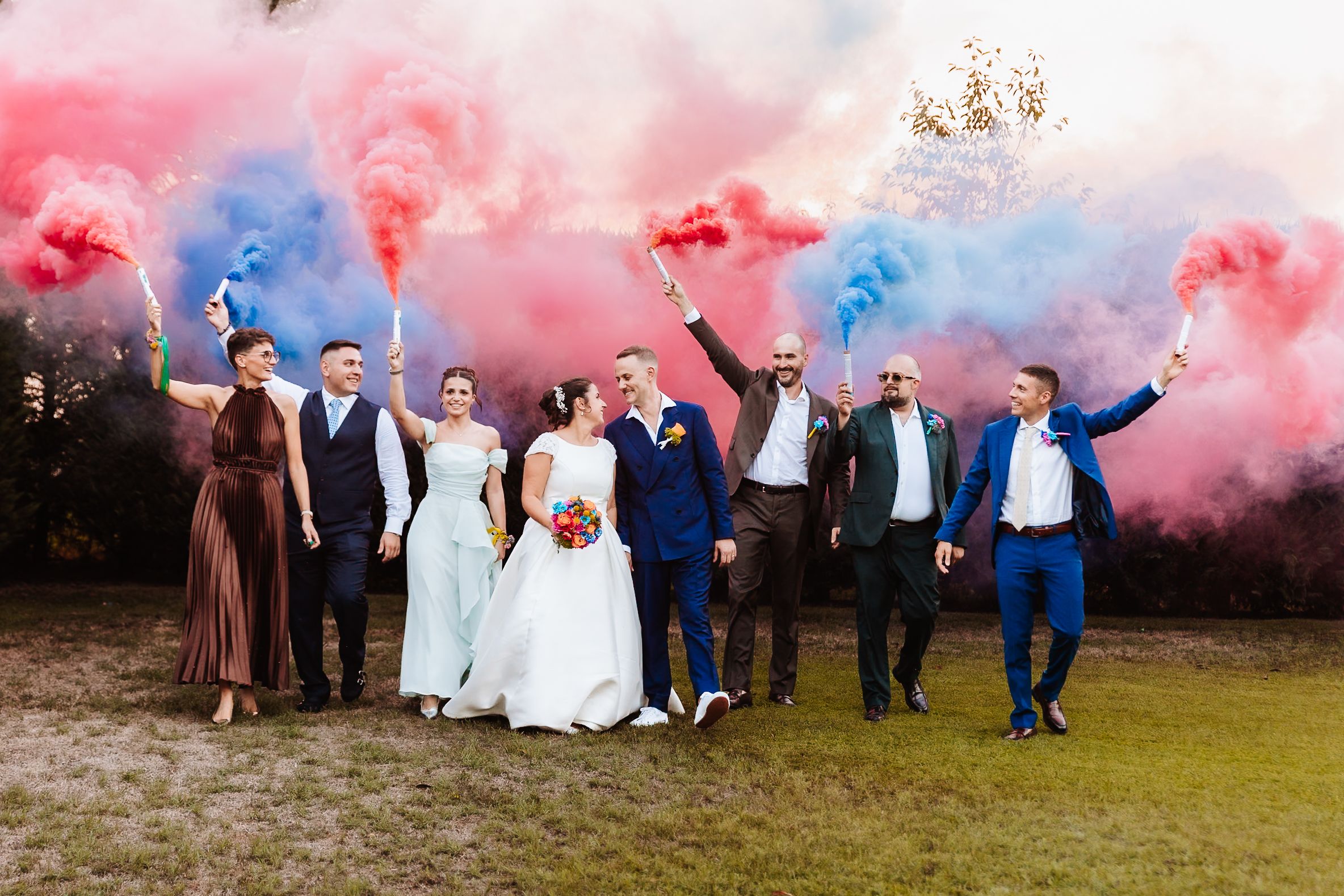 Wedding party celebrating outdoors with colorful red and blue smoke bombs, bride and groom surrounded by bridesmaids and groomsmen.