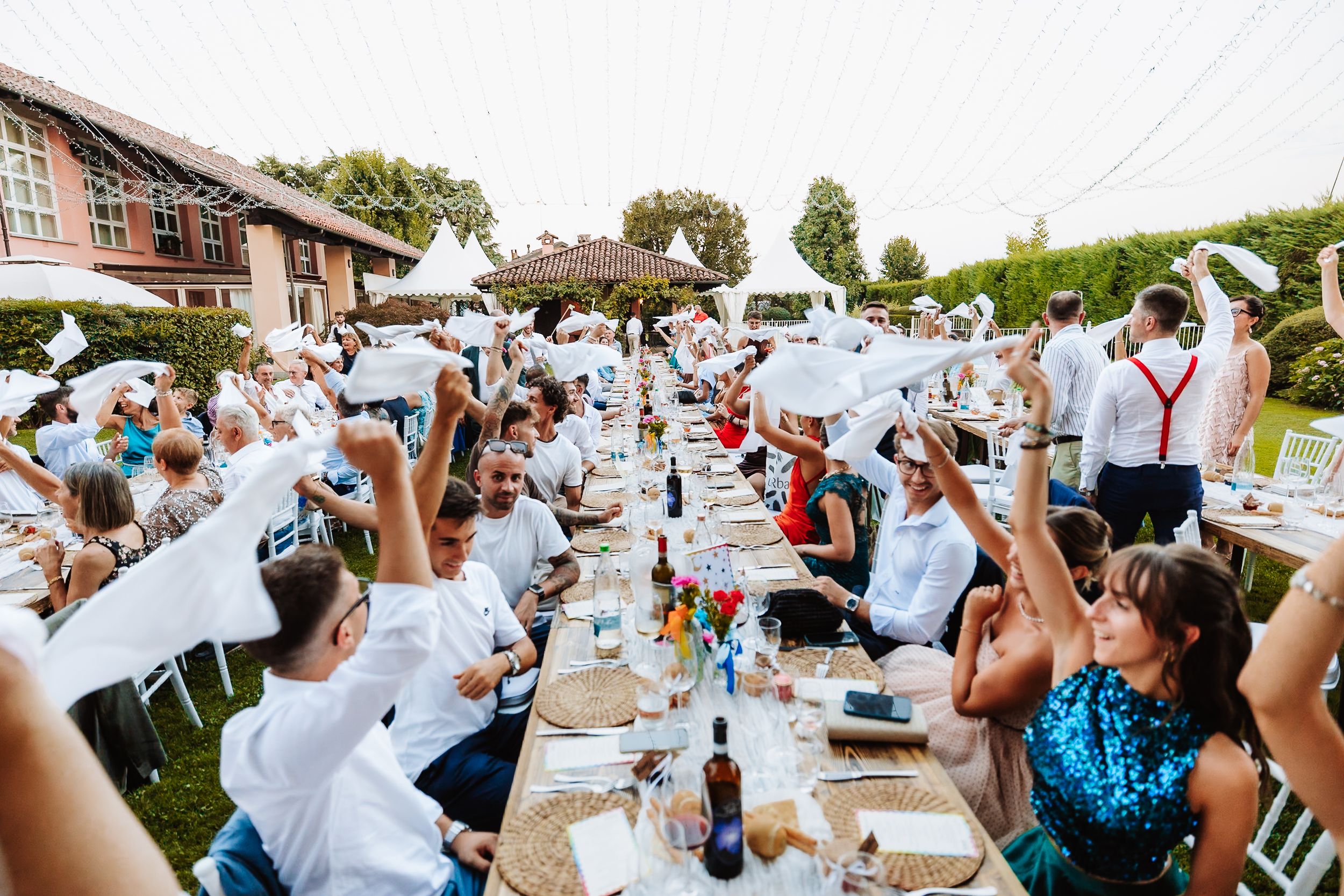 Joyful outdoor wedding reception with guests waving white napkins at long decorated tables in a garden setting.