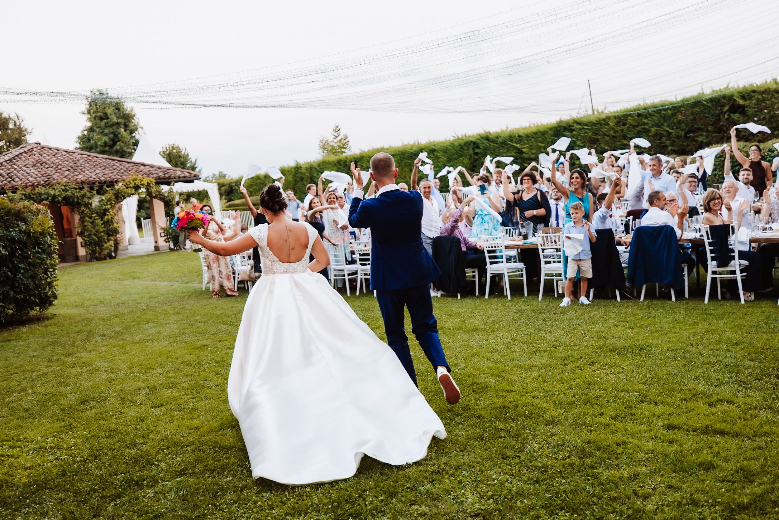 Bride and groom walk toward cheering guests waving white fans at an outdoor wedding reception.
