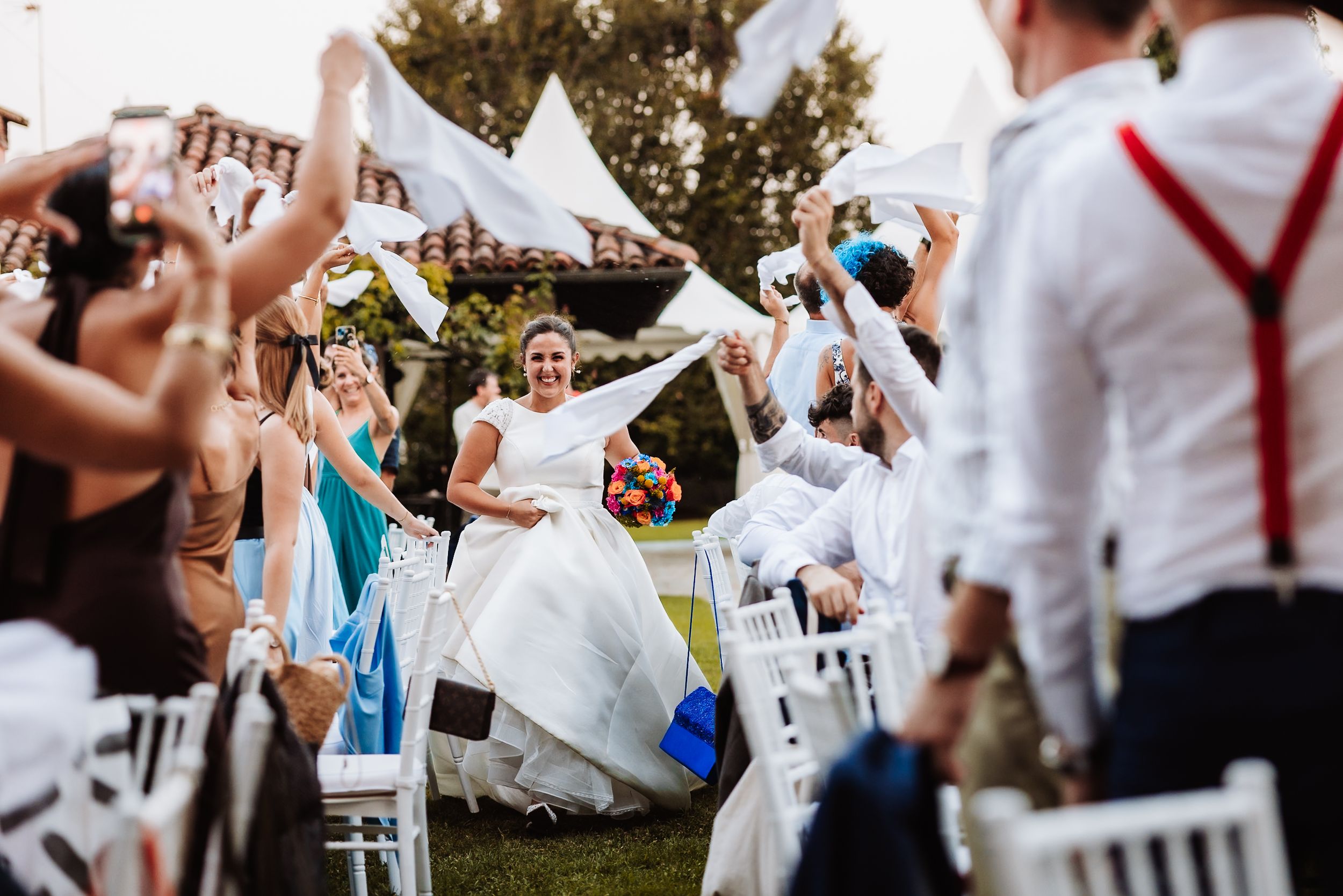 Joyful bride walks down outdoor wedding aisle as guests wave white paper fans in celebration.