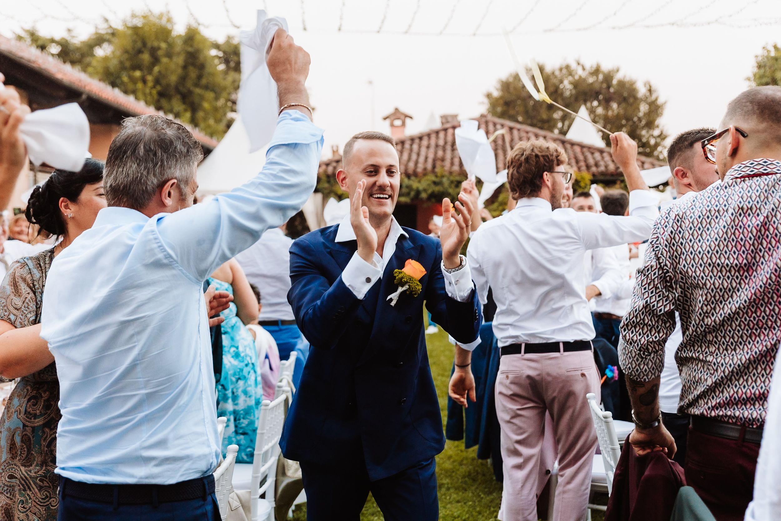 Smiling groom in navy suit walks through cheering wedding guests at outdoor ceremony.