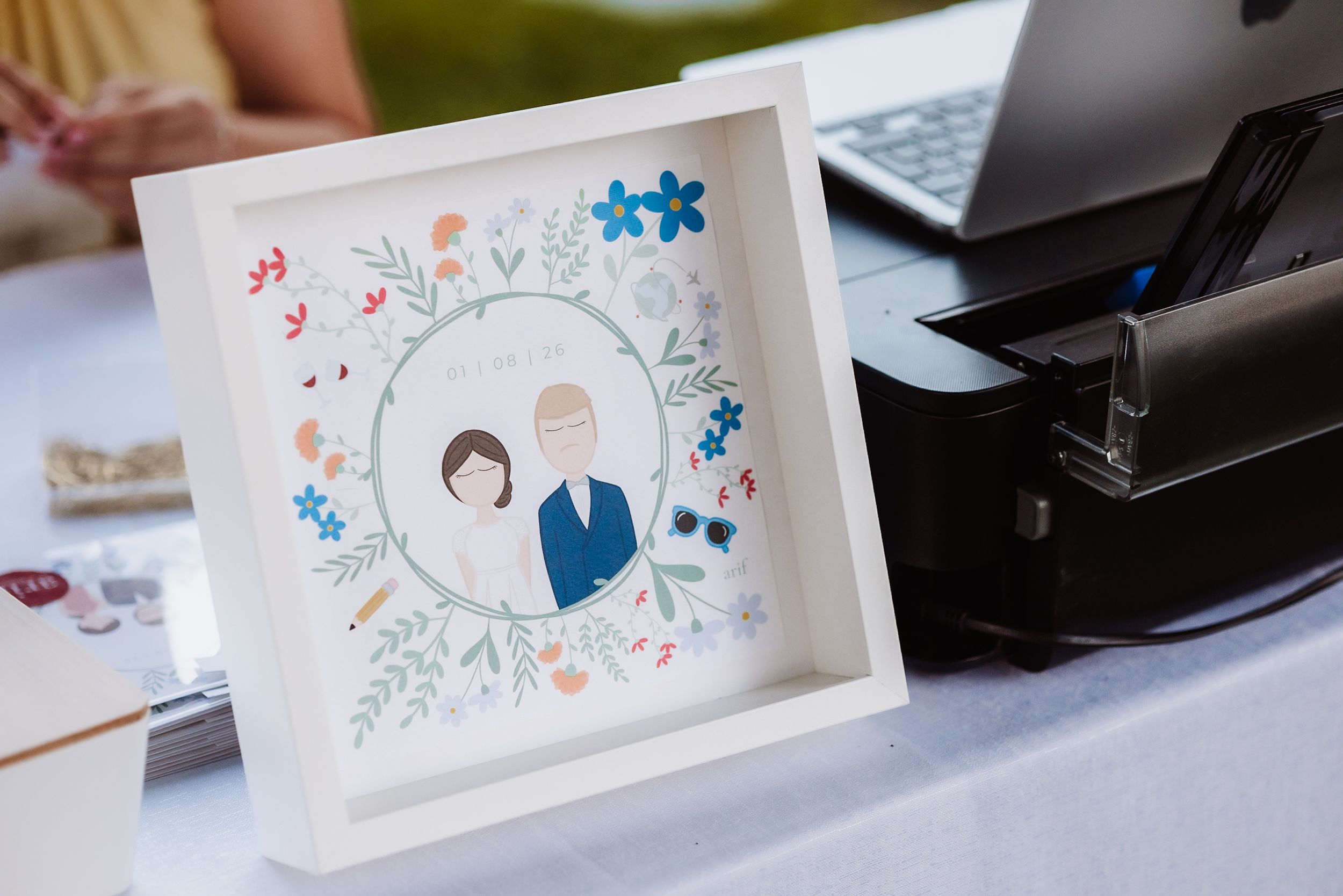 Framed illustrated portrait of bride and groom surrounded by colorful flowers displayed at wedding reception.
