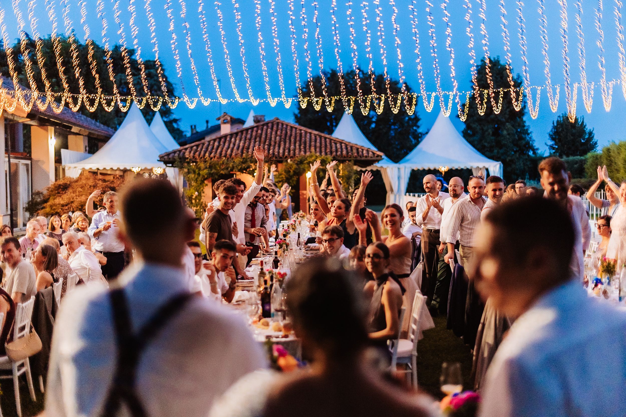 Wedding guests celebrate at an elegant outdoor reception under string lights at dusk.