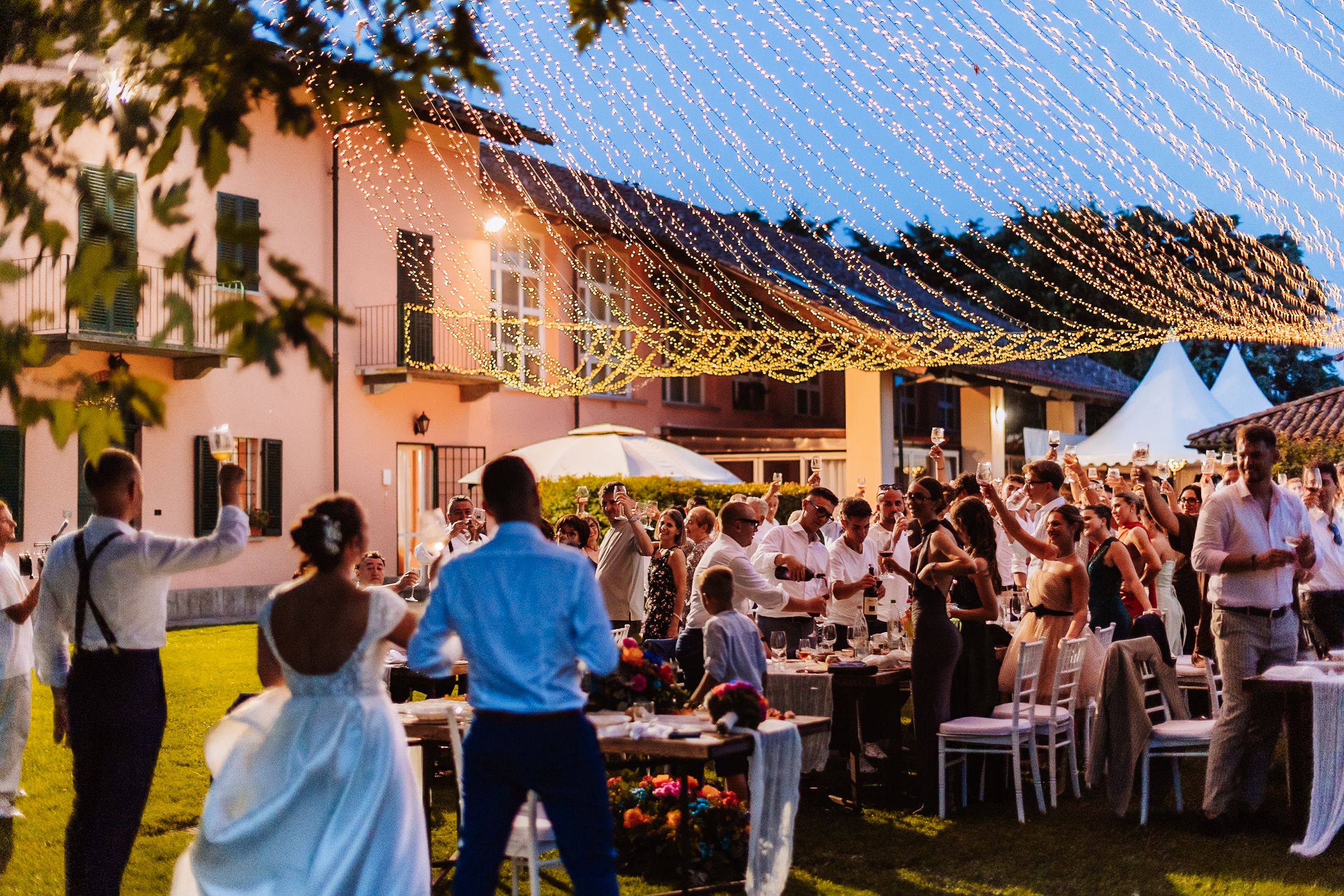 Elegant outdoor wedding reception at dusk with string lights overhead and guests celebrating around decorated tables.