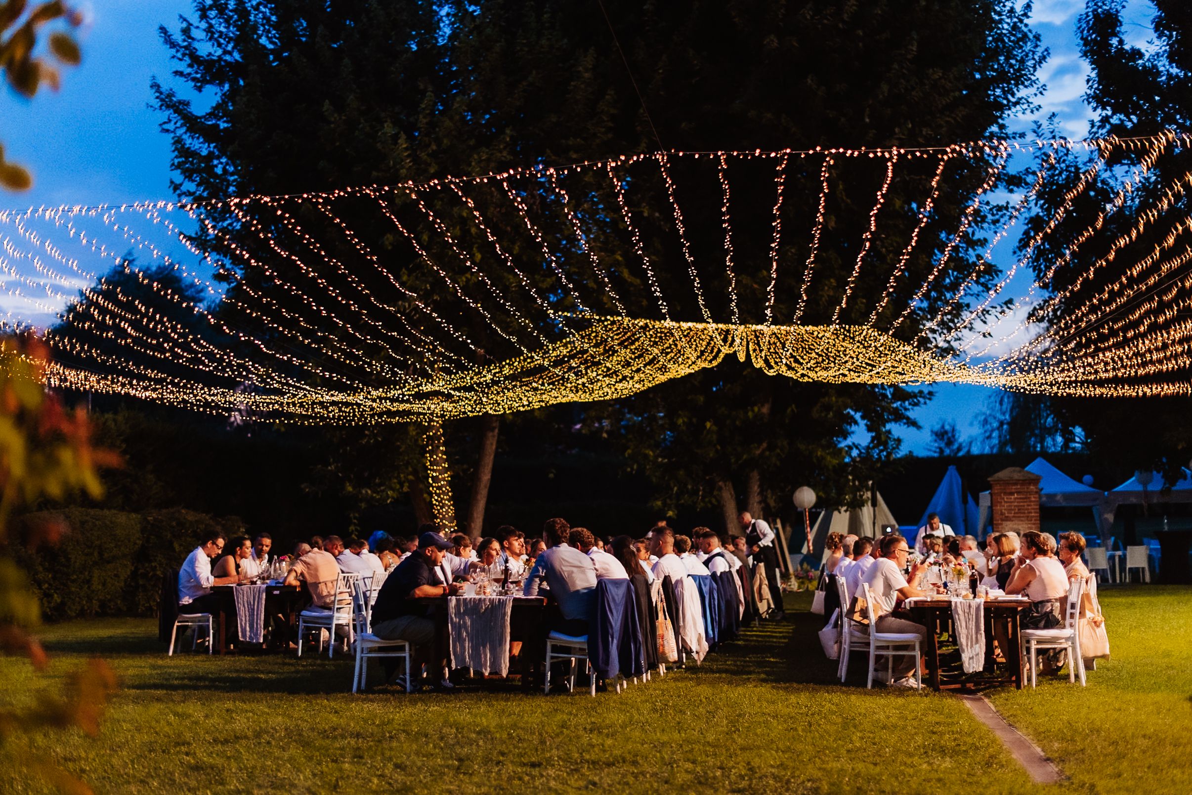 Romantic evening outdoor wedding reception under a canopy of fairy lights with guests dining on a lush green lawn.
