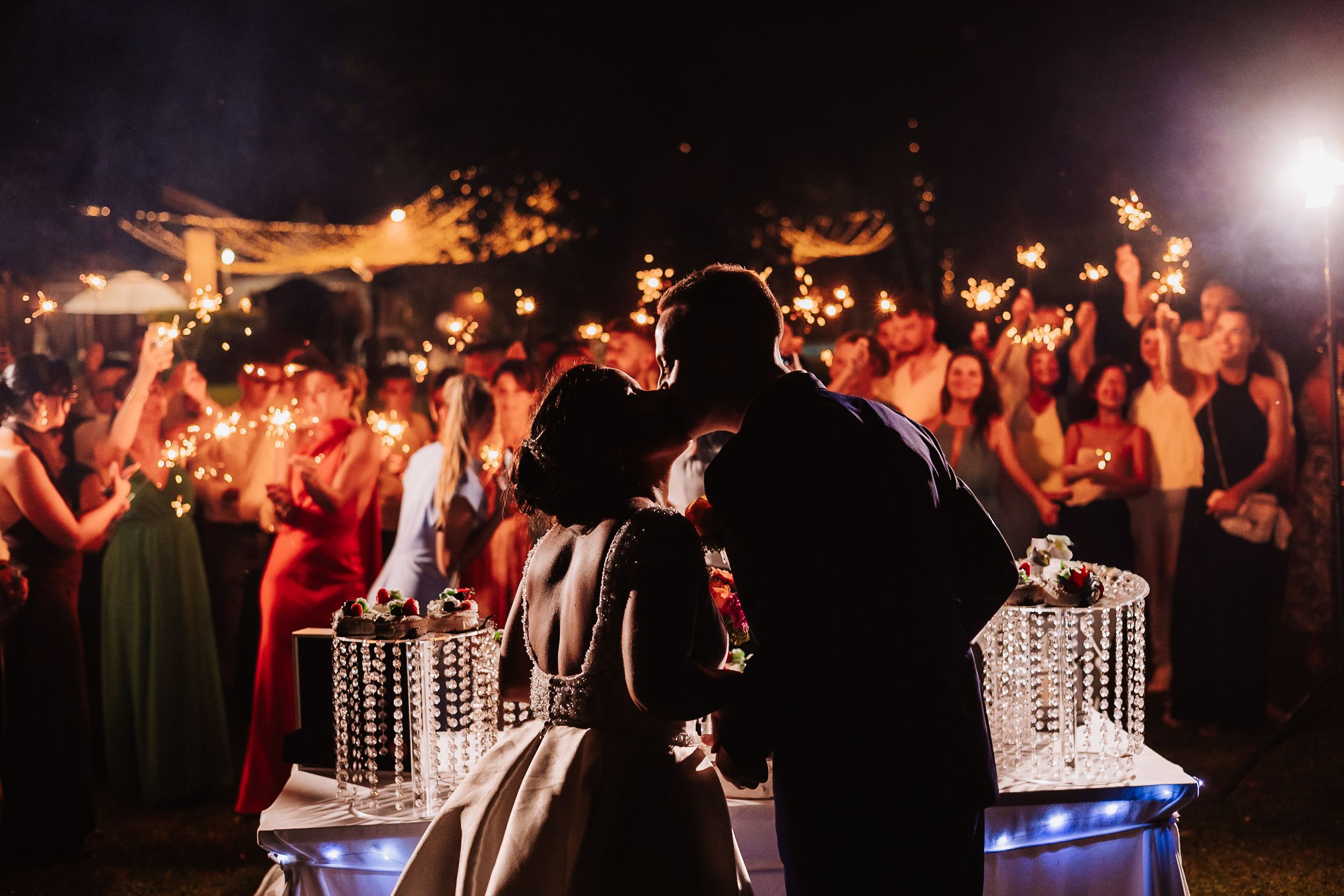 Bride and groom share a romantic kiss surrounded by sparkler-holding wedding guests at an outdoor evening reception.