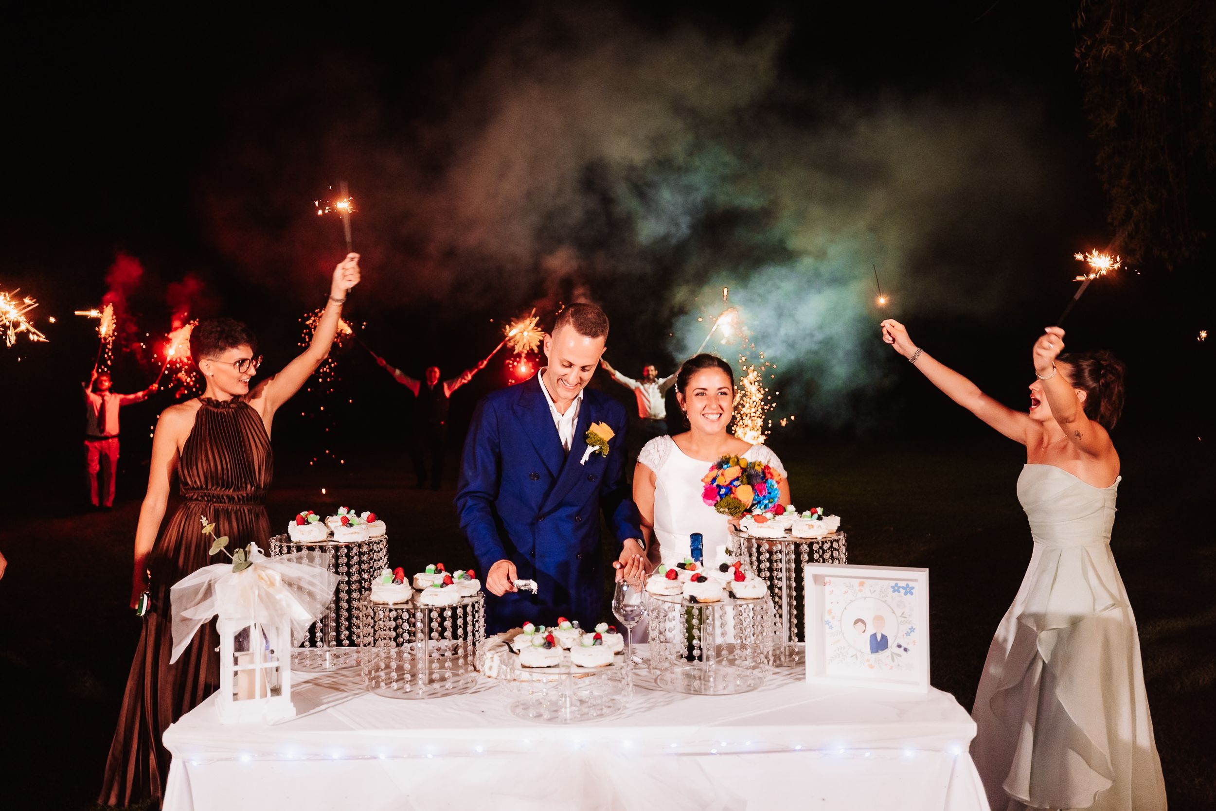 Bride and groom cut wedding cake surrounded by sparklers and guests at a nighttime celebration.