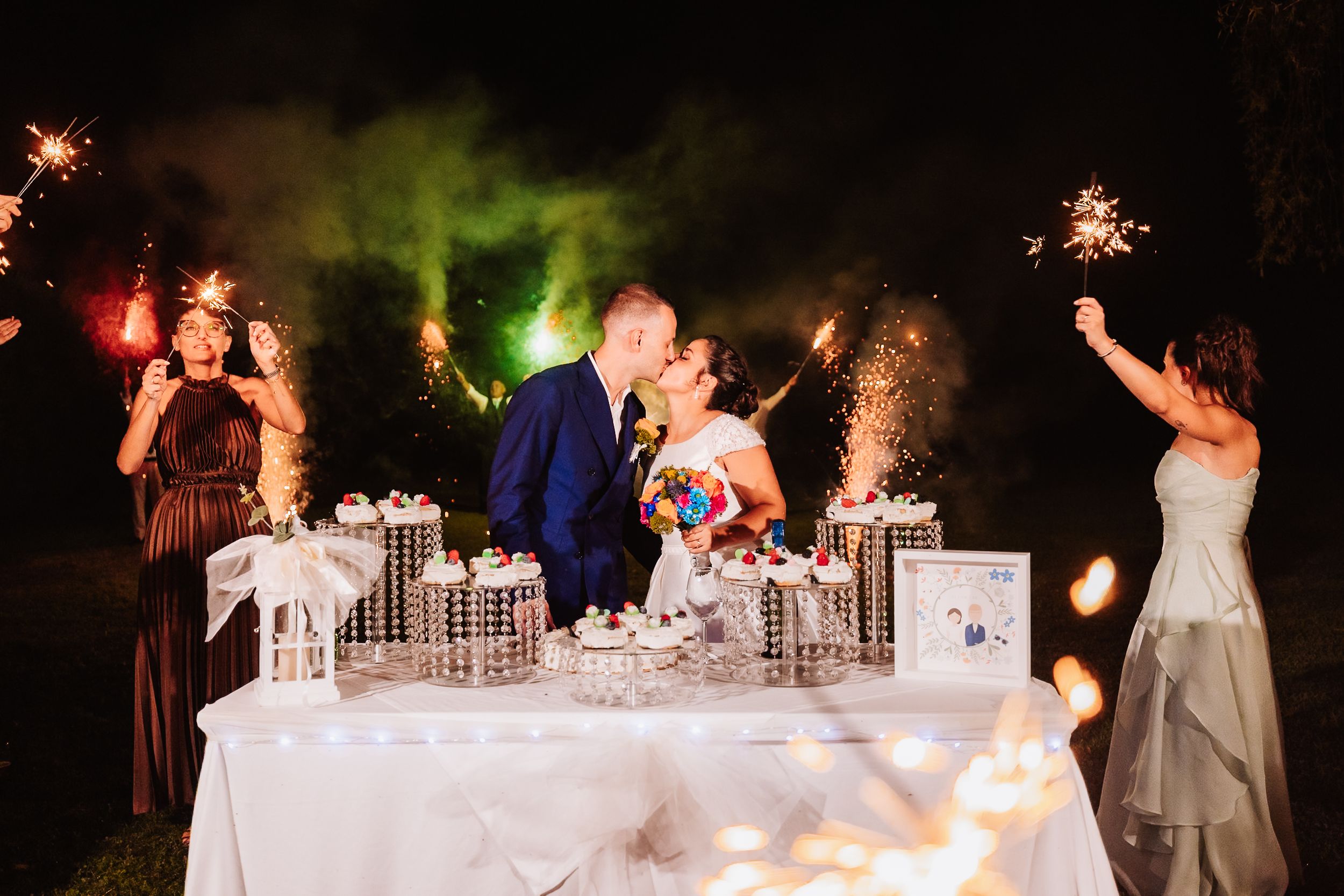 Bride and groom kiss at cake table surrounded by sparklers and bridesmaids at night.