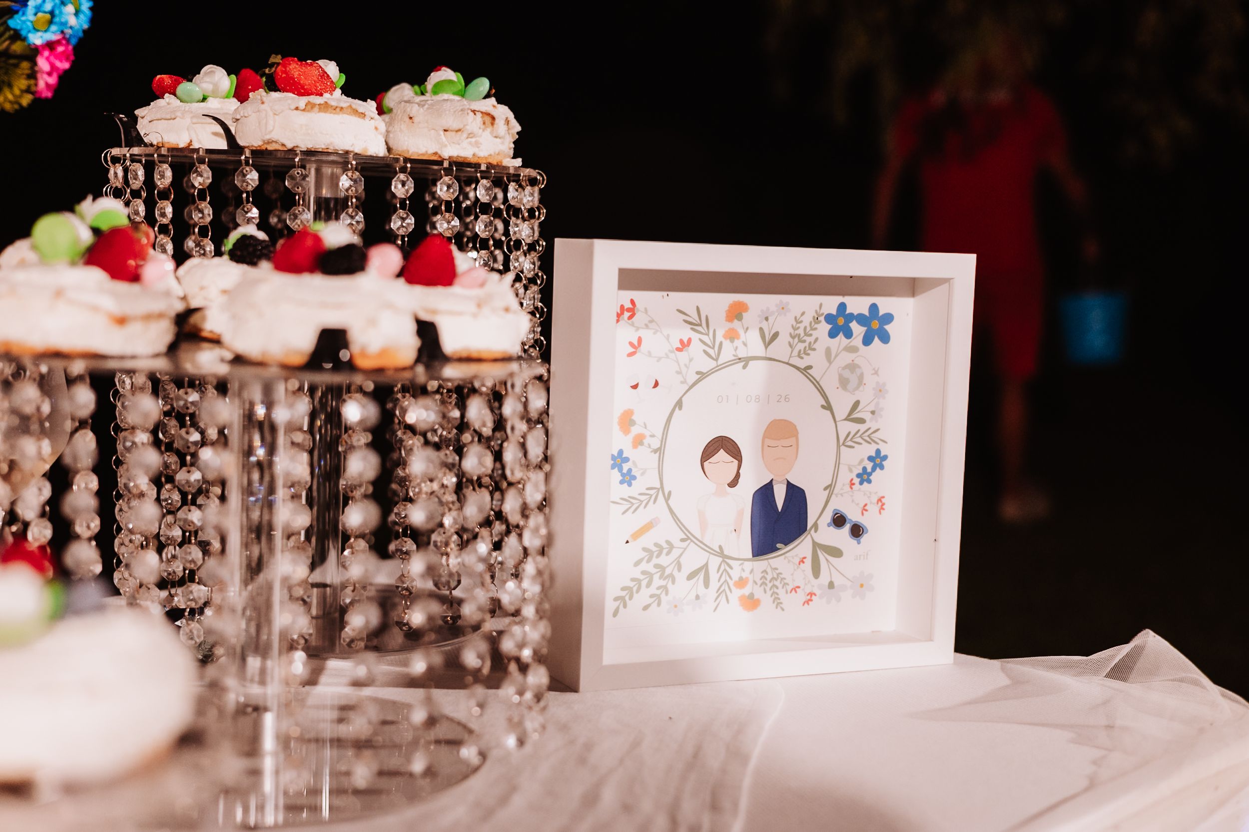 Wedding reception table with crystal cupcake stand and framed illustrated portrait of bride and groom surrounded by floral details.