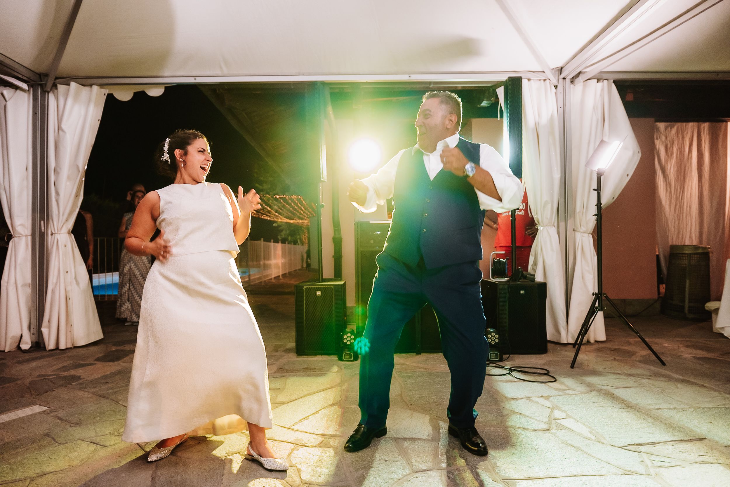 Bride and groom dancing joyfully at their wedding reception under a tent with bright stage lighting.