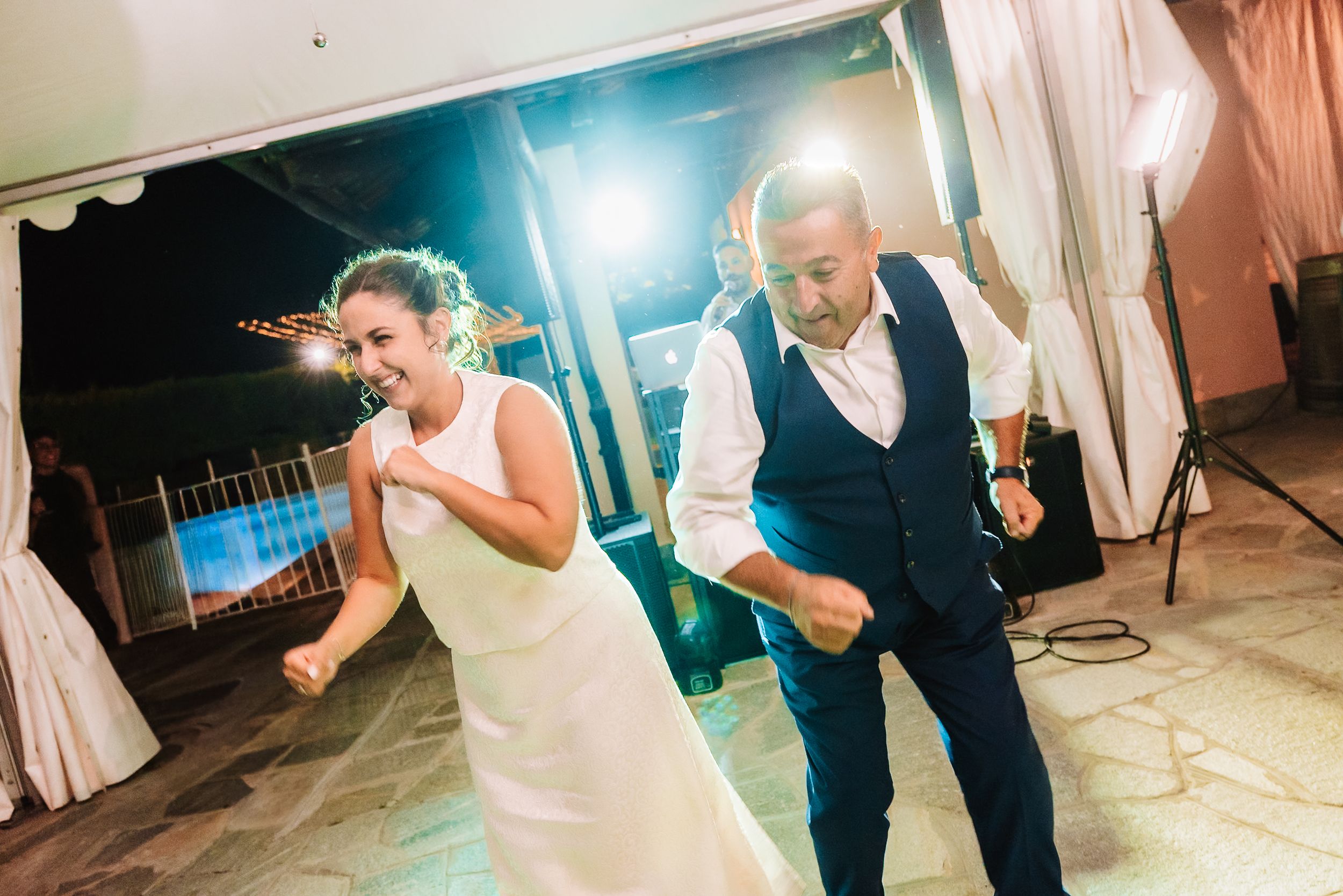 Bride and groom dancing joyfully at their outdoor wedding reception at night with bright lights in the background.