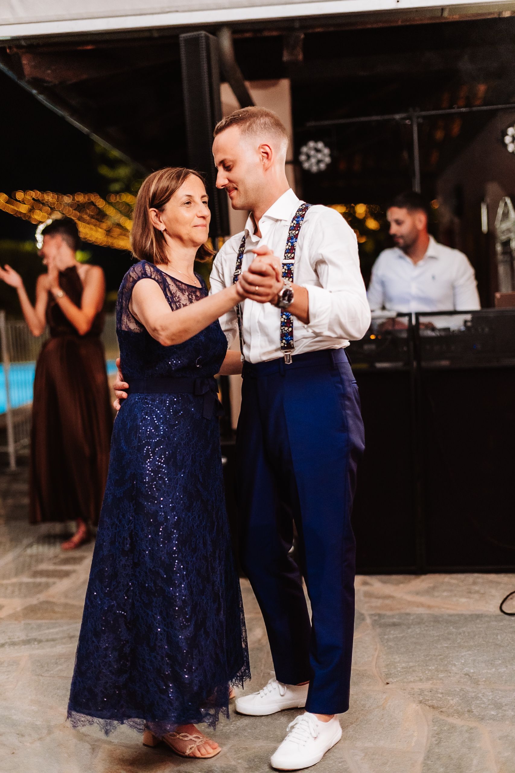 Groom in navy suspenders shares a mother-son dance at an outdoor wedding reception with string lights.