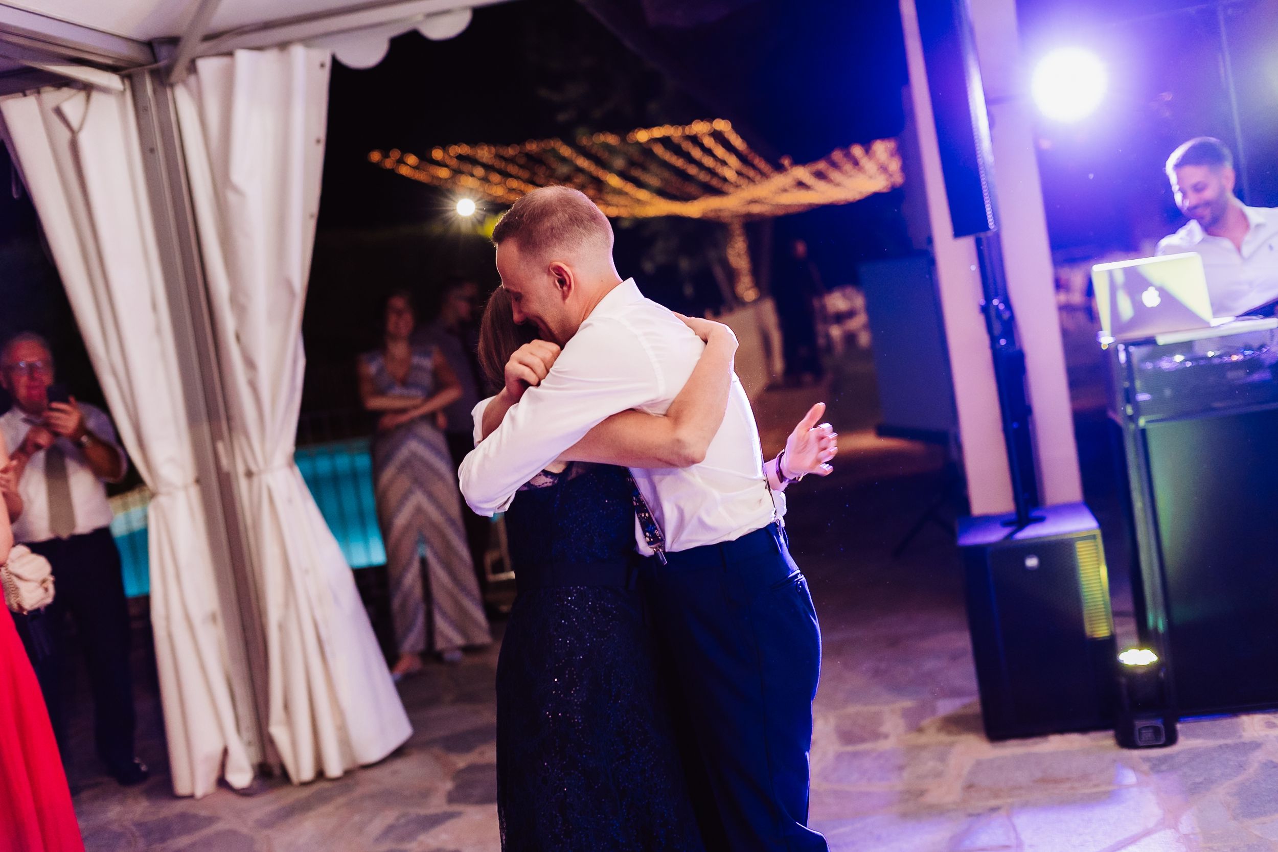 Two grooms share an emotional first dance embrace at an outdoor wedding reception with string lights.