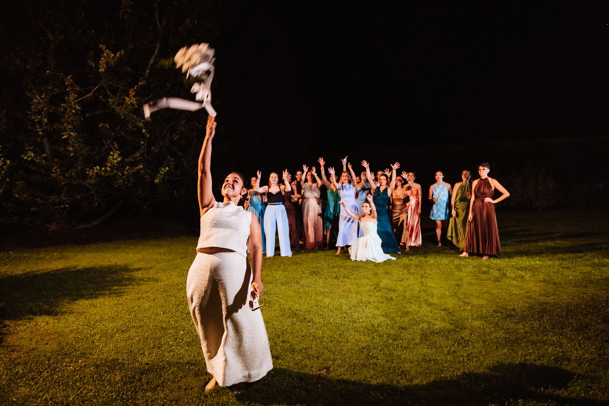 Bride tosses bouquet to excited wedding guests gathered on green lawn at night during outdoor reception celebration.