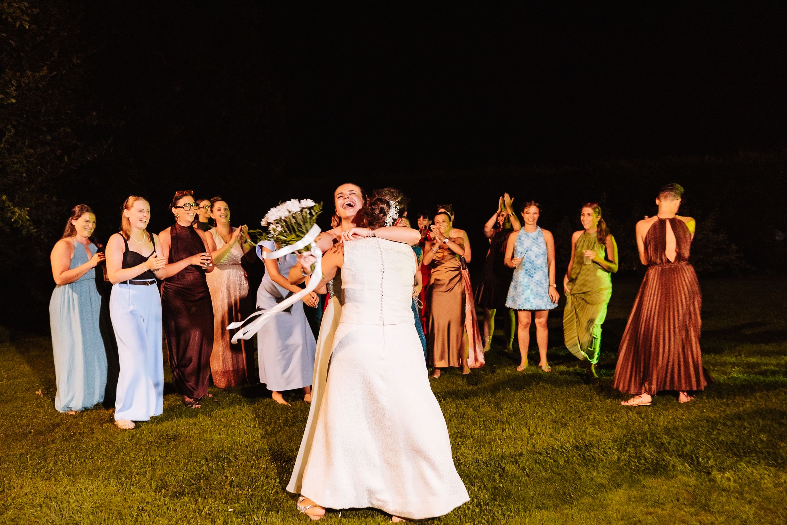 Bride tossing bouquet to excited bridesmaids in colorful dresses during outdoor nighttime wedding celebration.