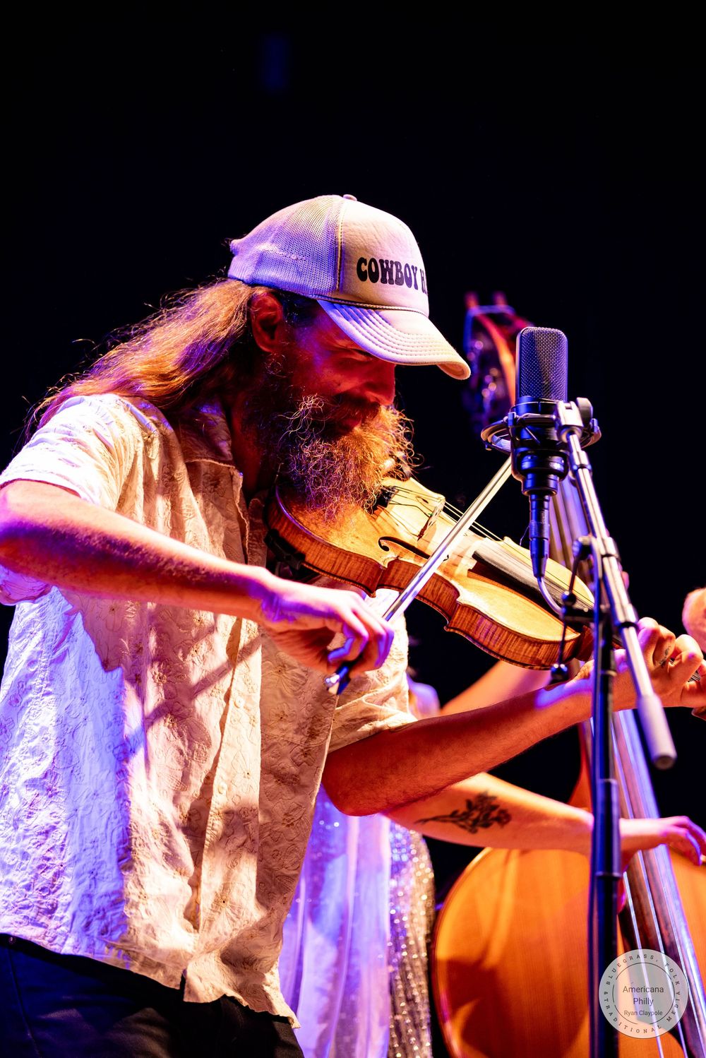 Bearded musician with long hair and cowboy hat plays violin on stage under purple lights.