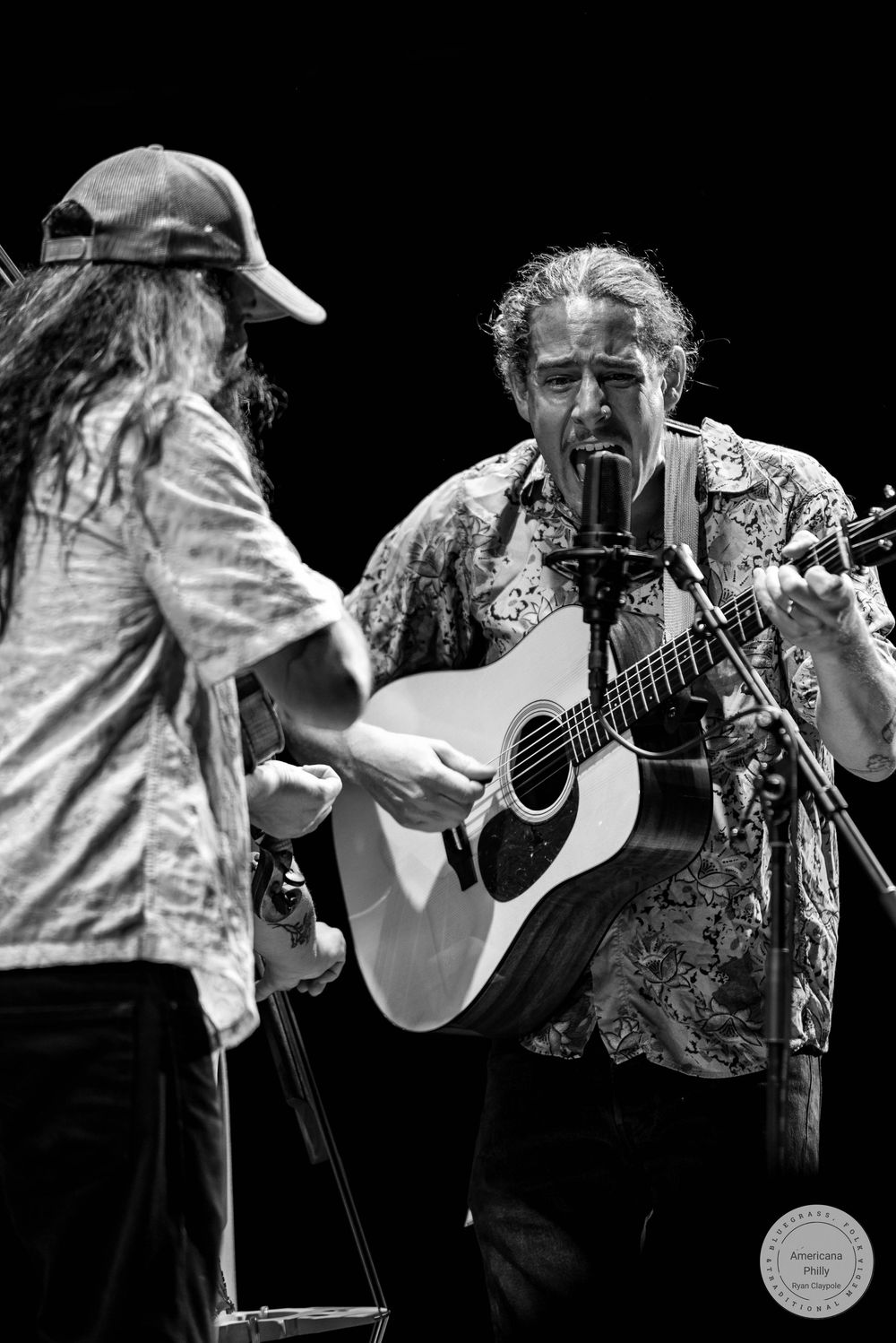 Two musicians perform on stage, one playing acoustic guitar at a microphone in black and white concert photography.