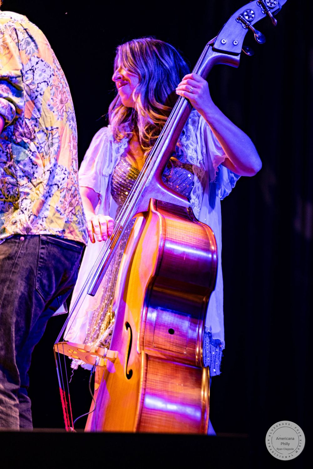 Woman playing upright bass on stage under purple and orange stage lights wearing white flowing top.