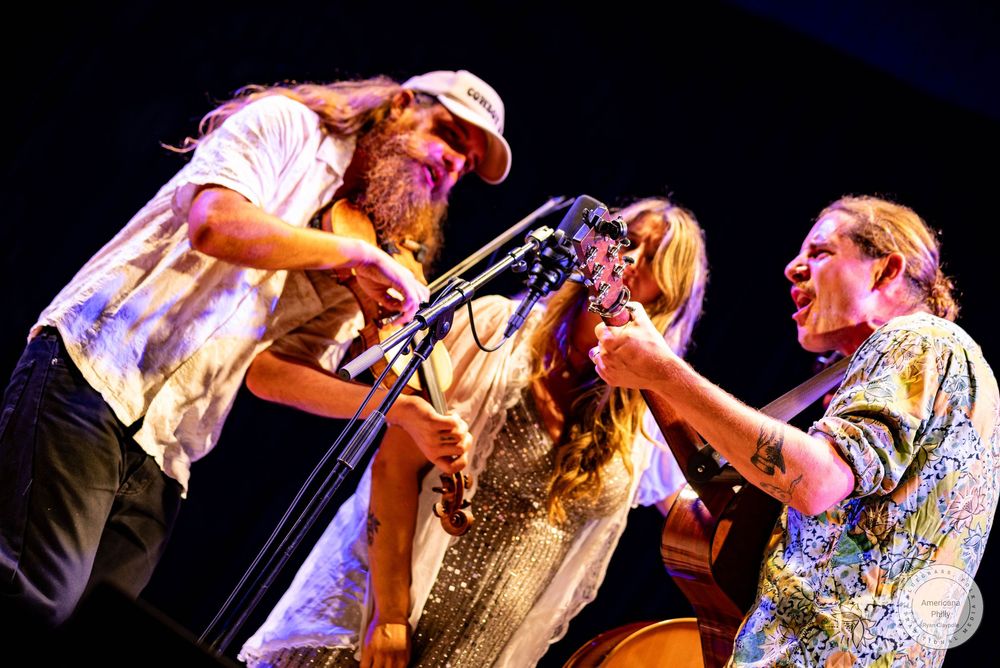 Three musicians share a microphone on stage, playing guitars and singing together under bright concert lights.