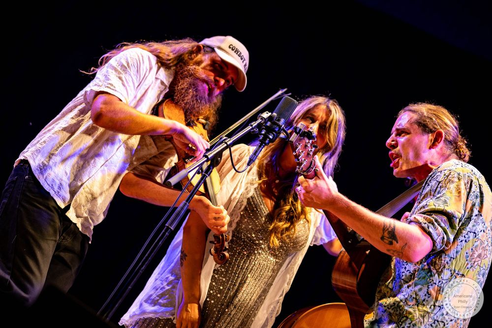 Three musicians performing on stage, two men and a woman playing guitars and violin under warm stage lights.