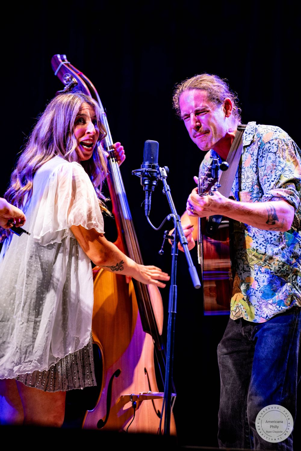 Two musicians perform on stage, a woman playing upright bass and a man playing guitar, both smiling joyfully.