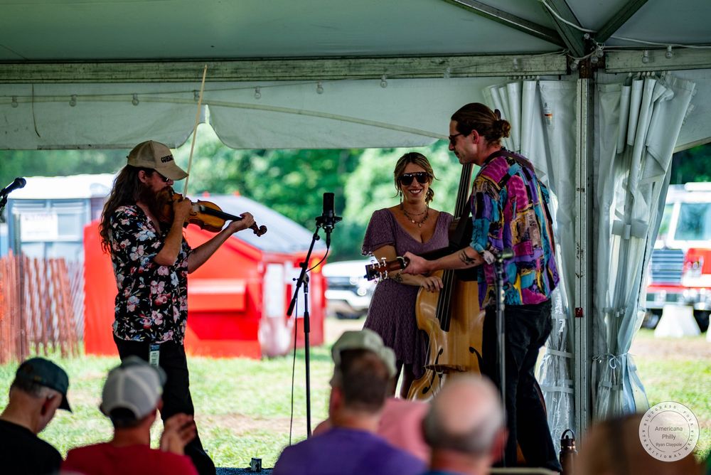 Three musicians perform under a tent outdoors, playing violin and guitar at a folk music festival.