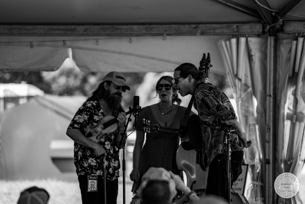 Black and white photo of three musicians performing under a tent at an outdoor music festival.